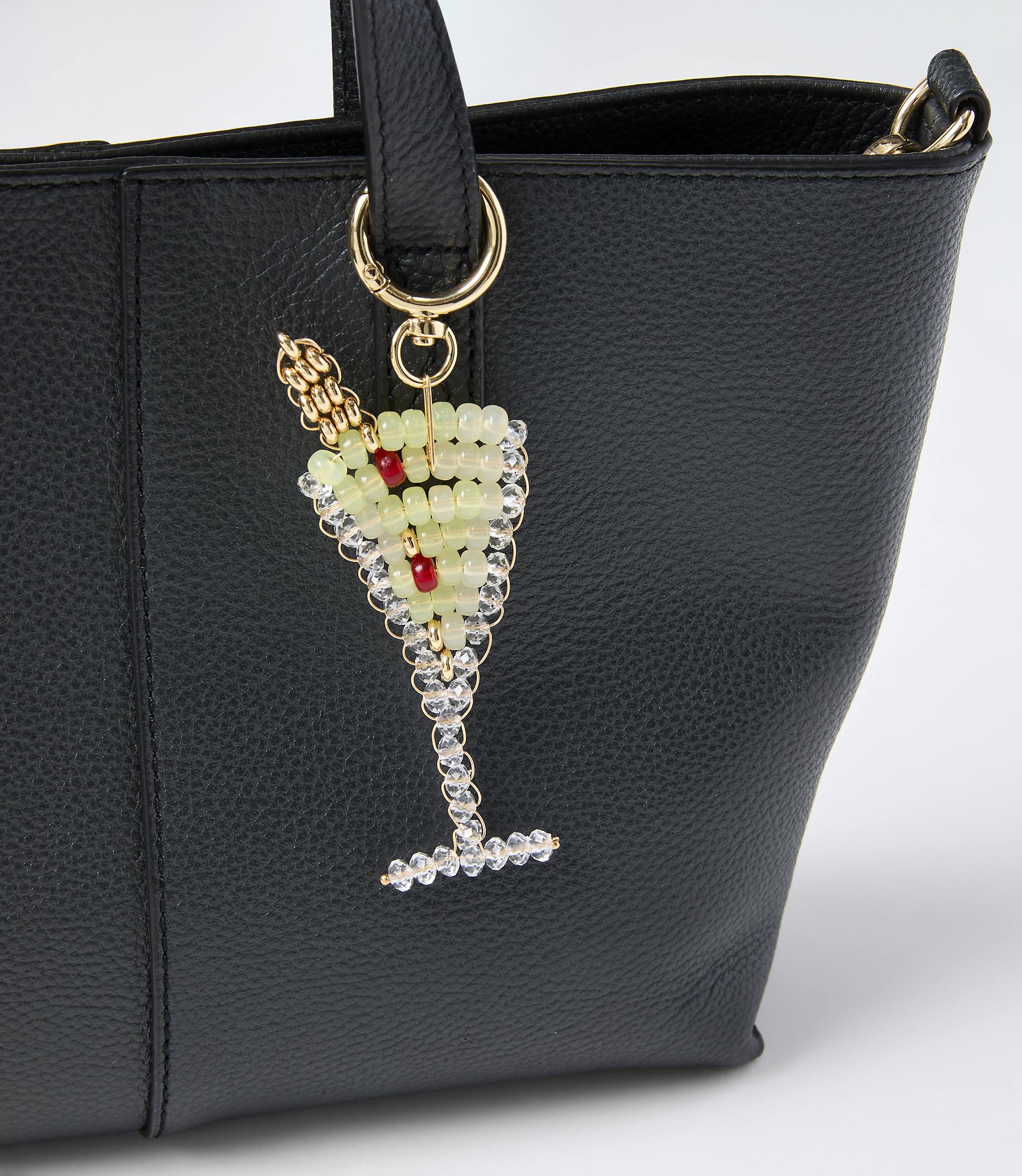 A beaded champagne glass charm hangs from the handle of a black leather handbag against a plain white background.