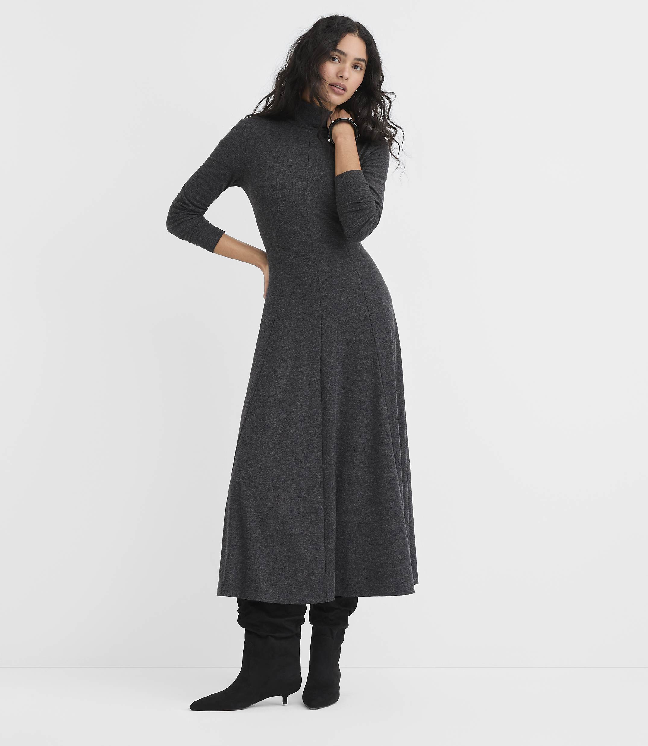 A woman wearing a long-sleeved charcoal grey maxi dress and black boots stands against a plain white background.