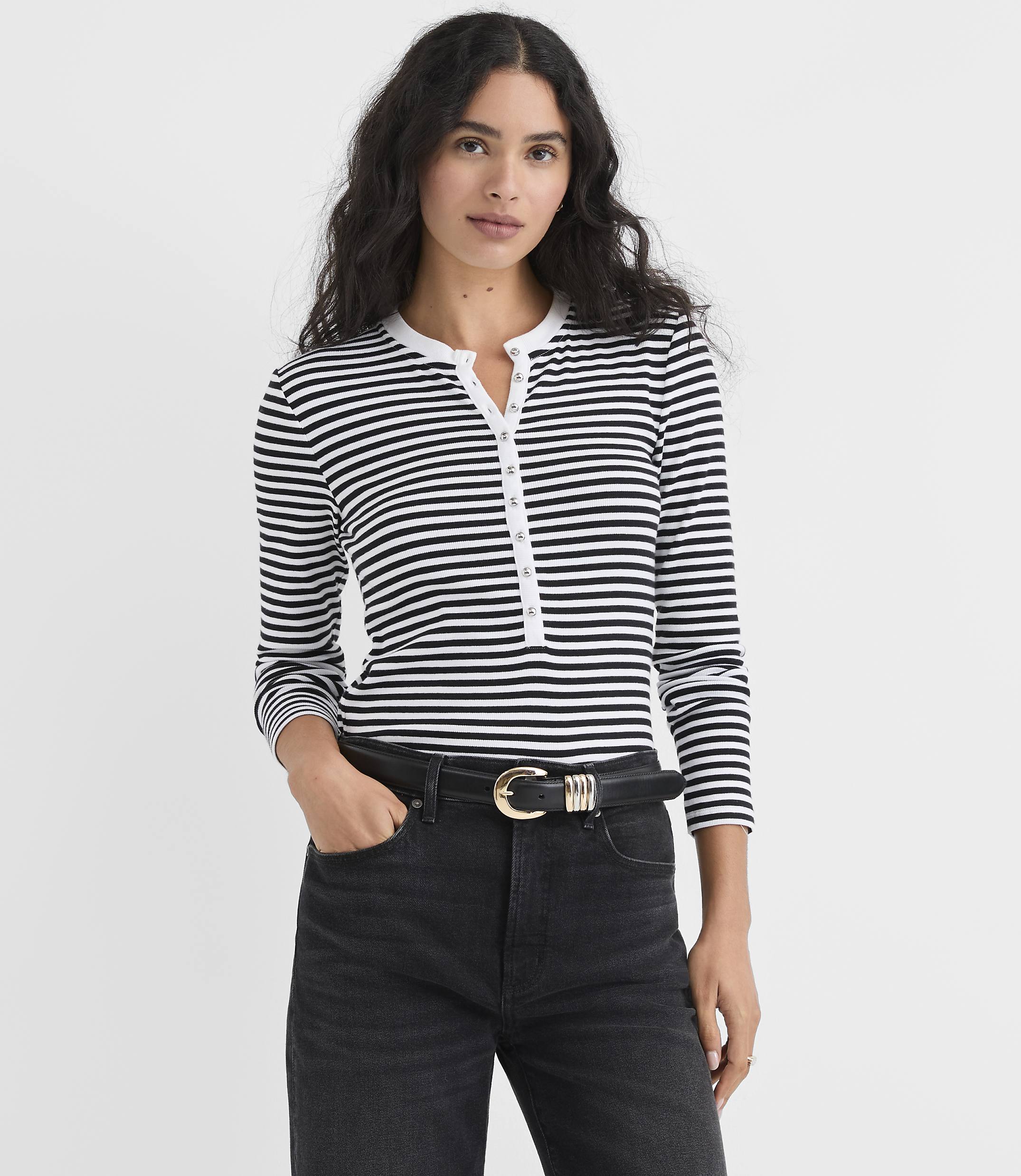 A woman wearing a black and white striped long-sleeve shirt and black jeans stands against a plain white background.