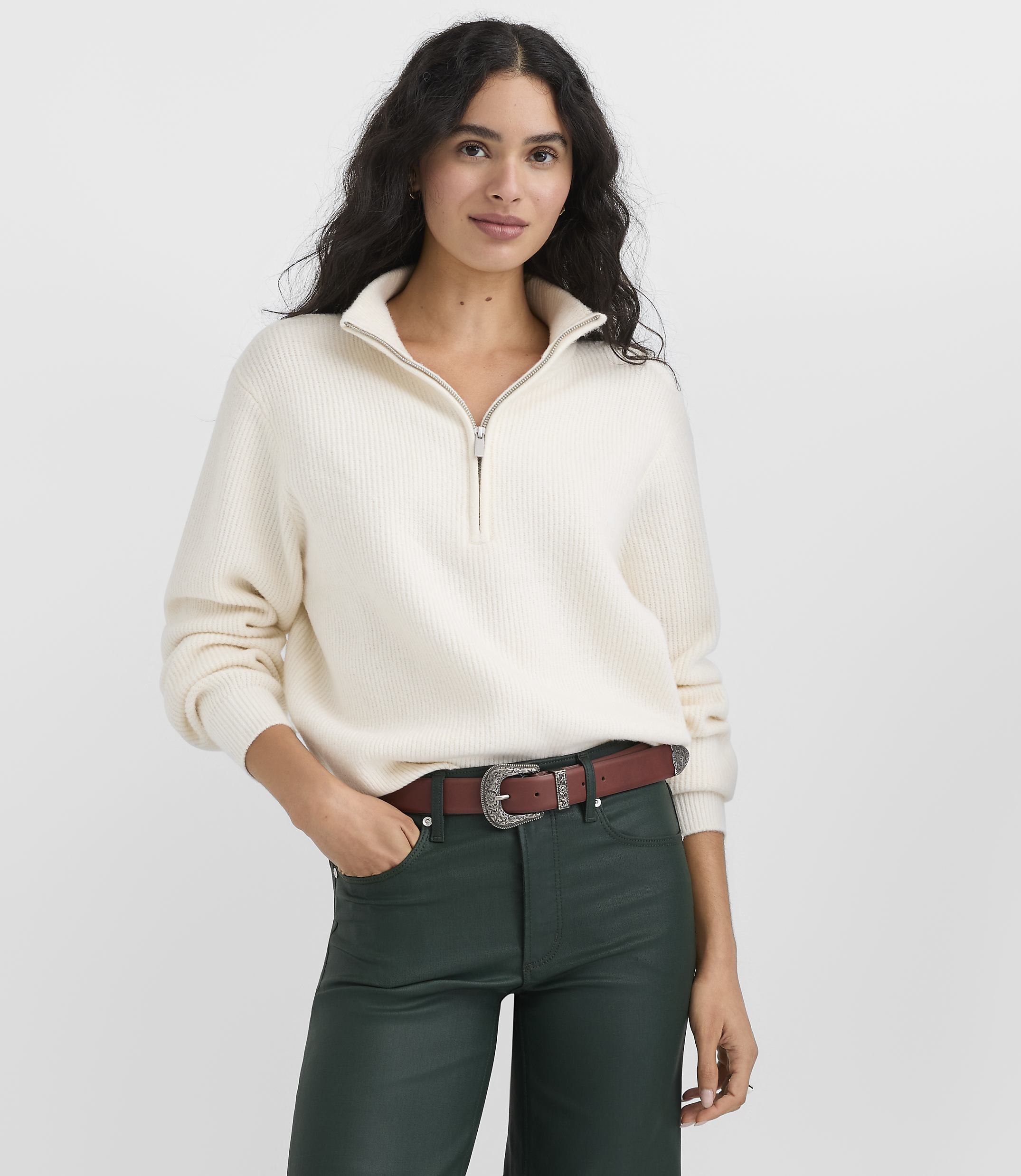 A woman wearing a cream-colored quarter-zip sweater and dark green leather pants stands against a plain light gray background.