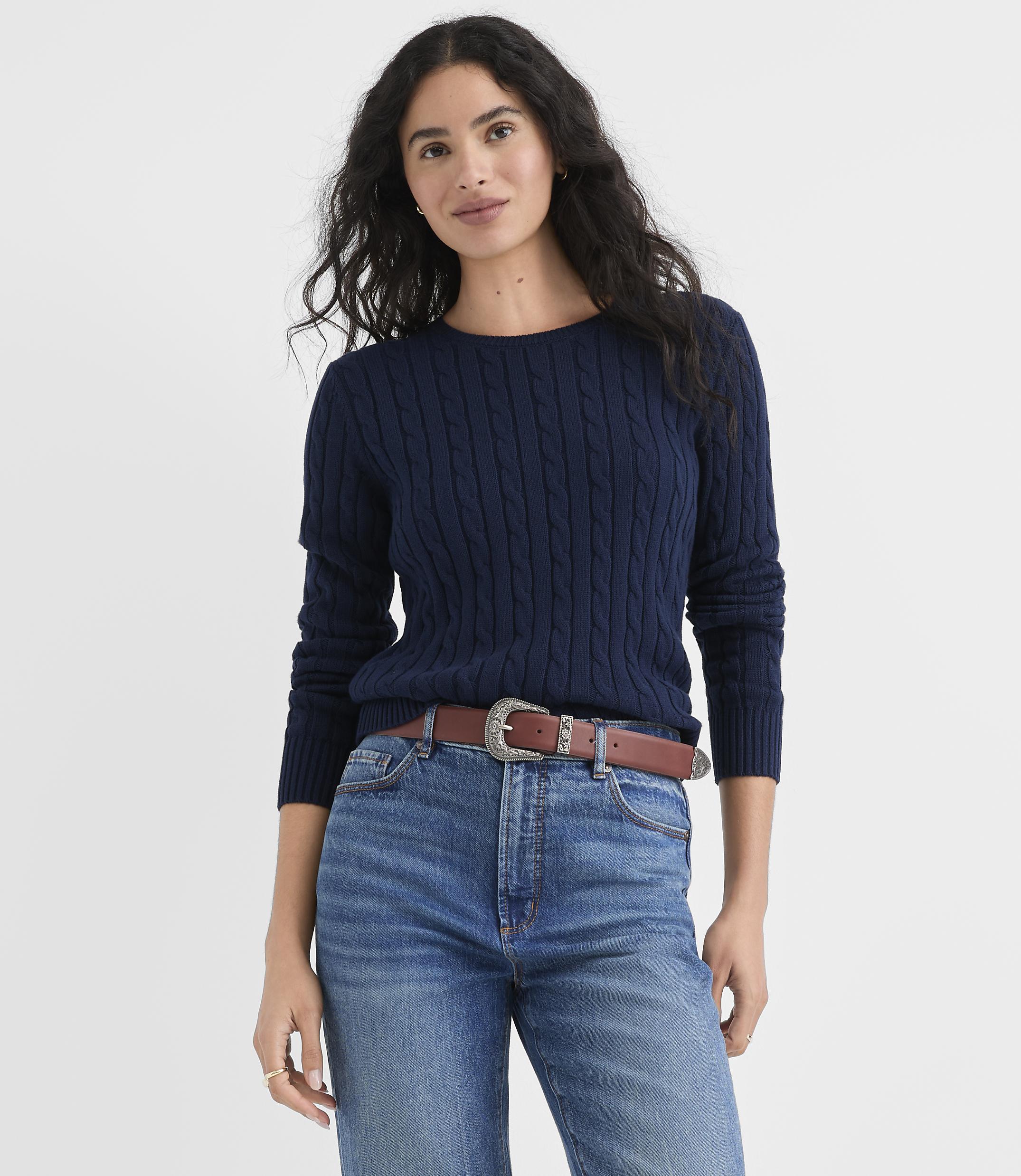 A woman wearing a navy cable-knit sweater, blue jeans, and a brown belt stands against a plain white background.