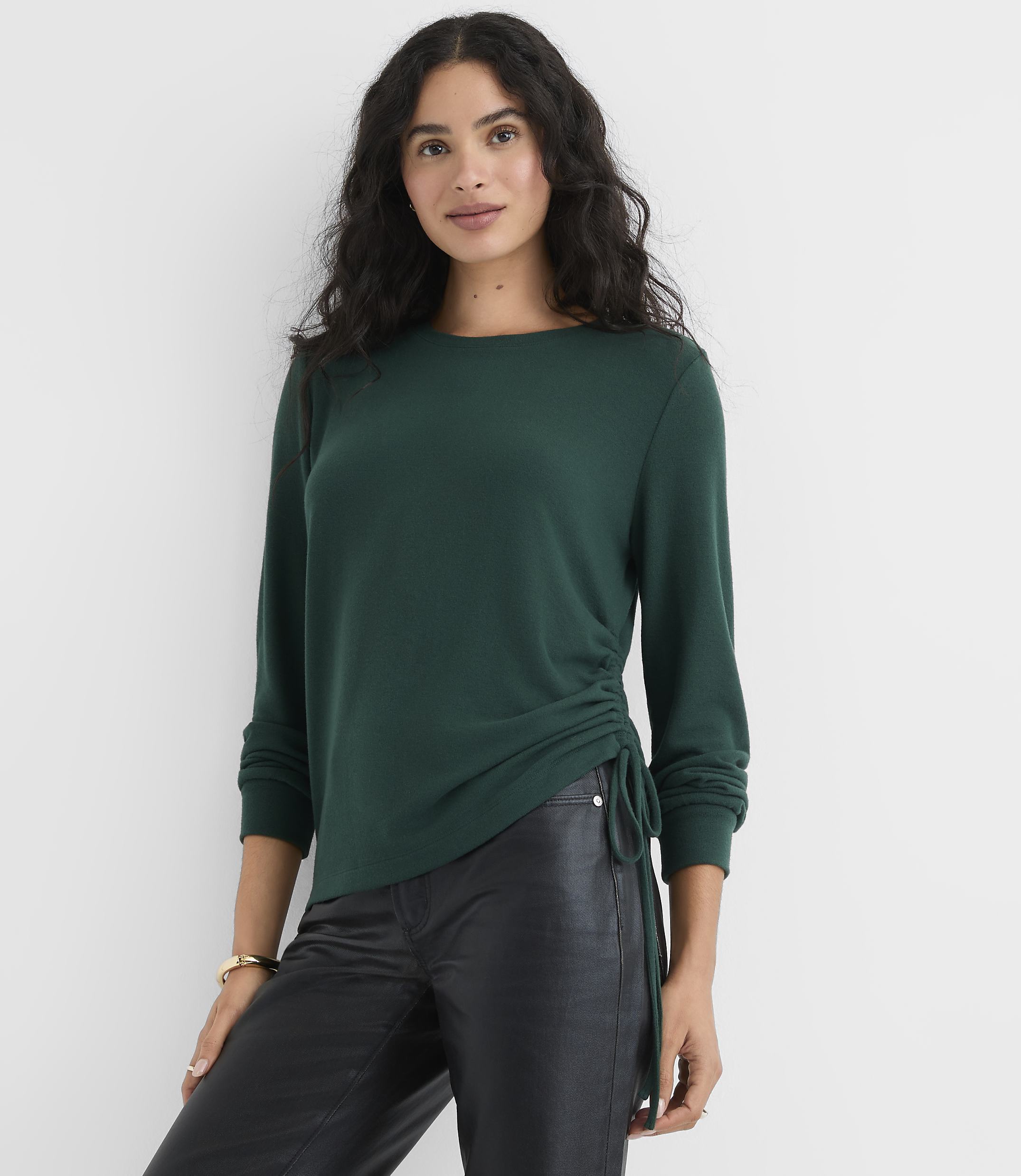 A woman wearing a dark green long-sleeved top and black leather pants stands against a plain white background.