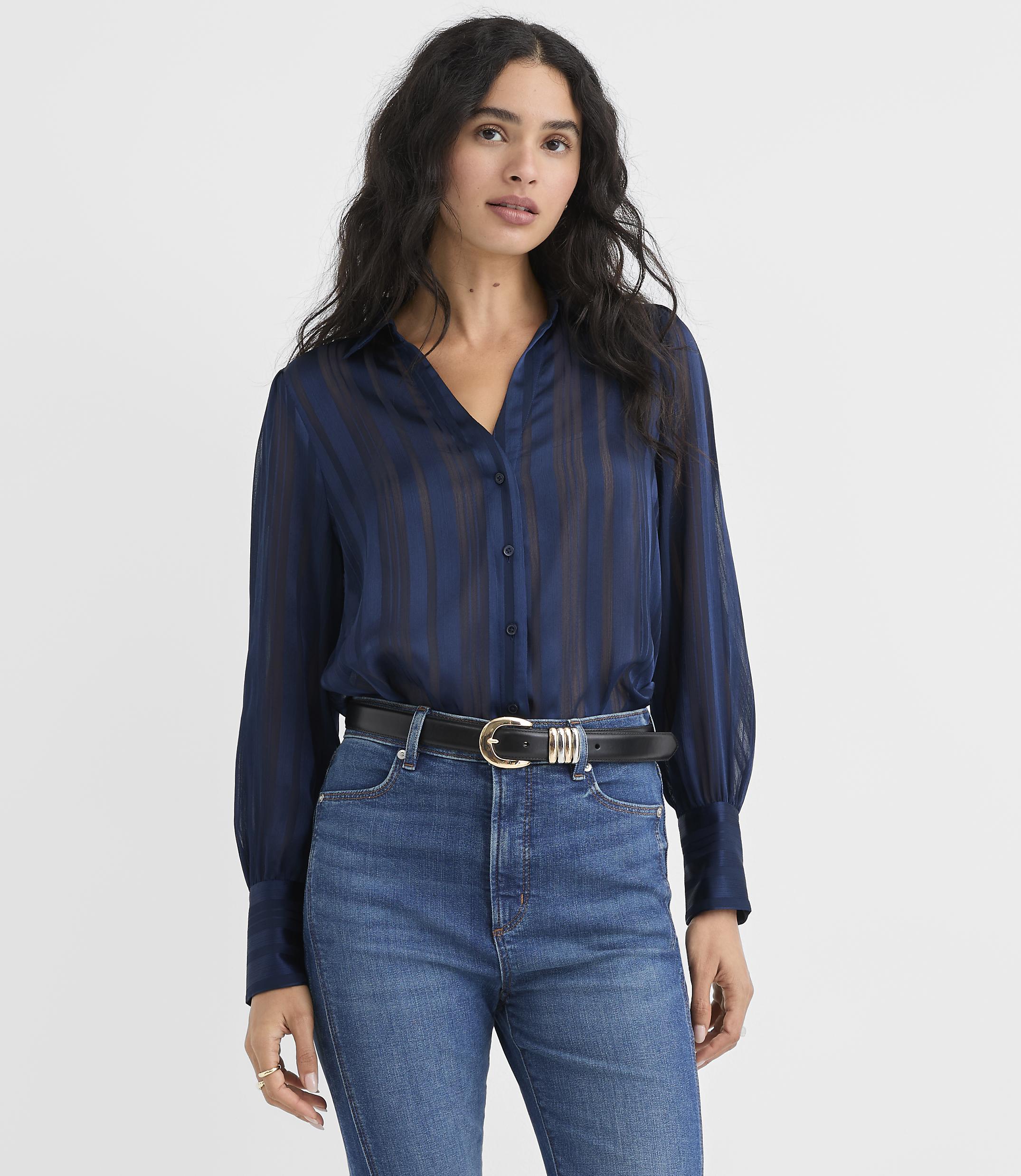 A woman wearing a navy blue striped blouse and blue jeans stands against a plain white background.