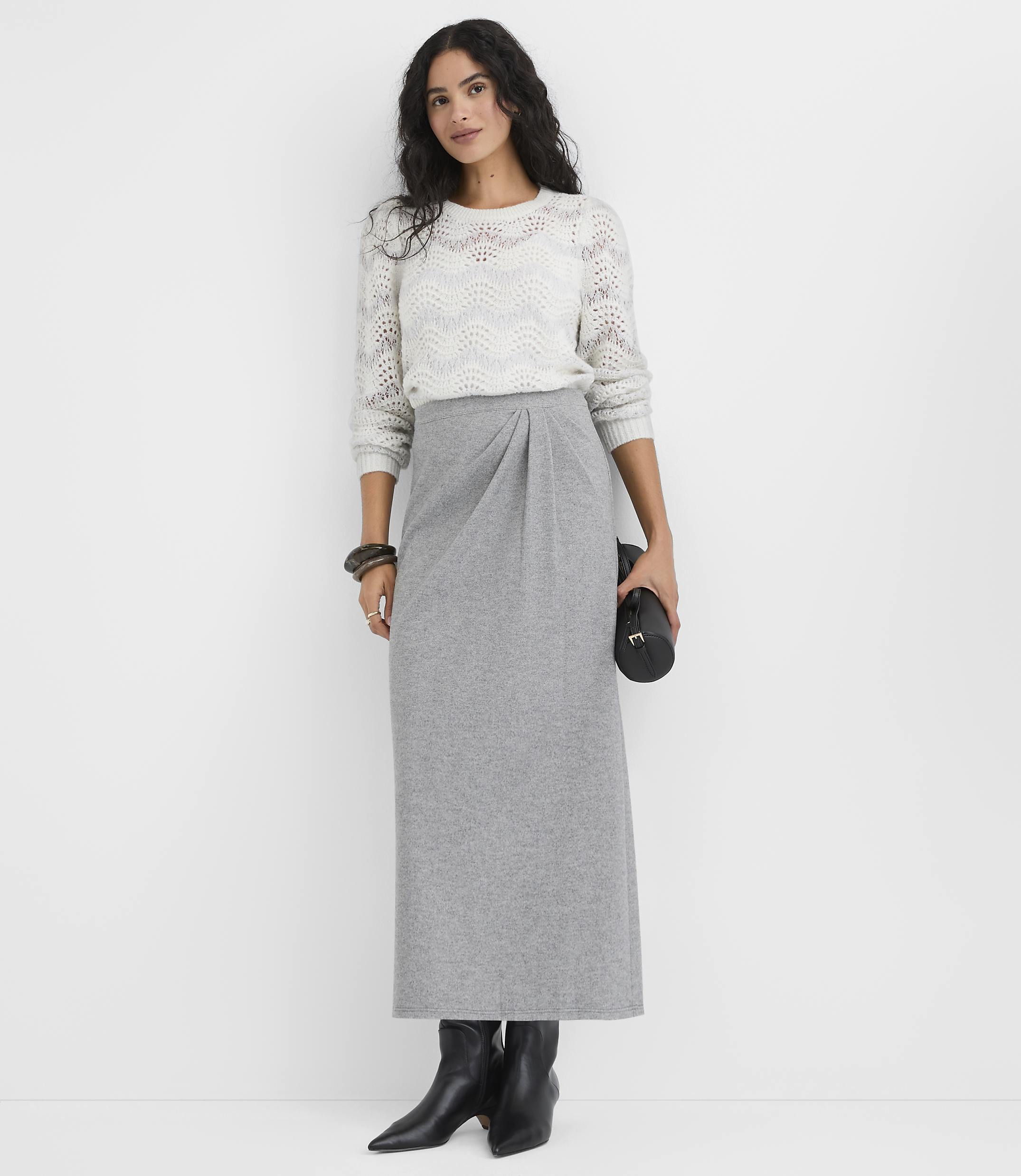 A woman wearing a white knit sweater and a long grey skirt stands against a plain white background.