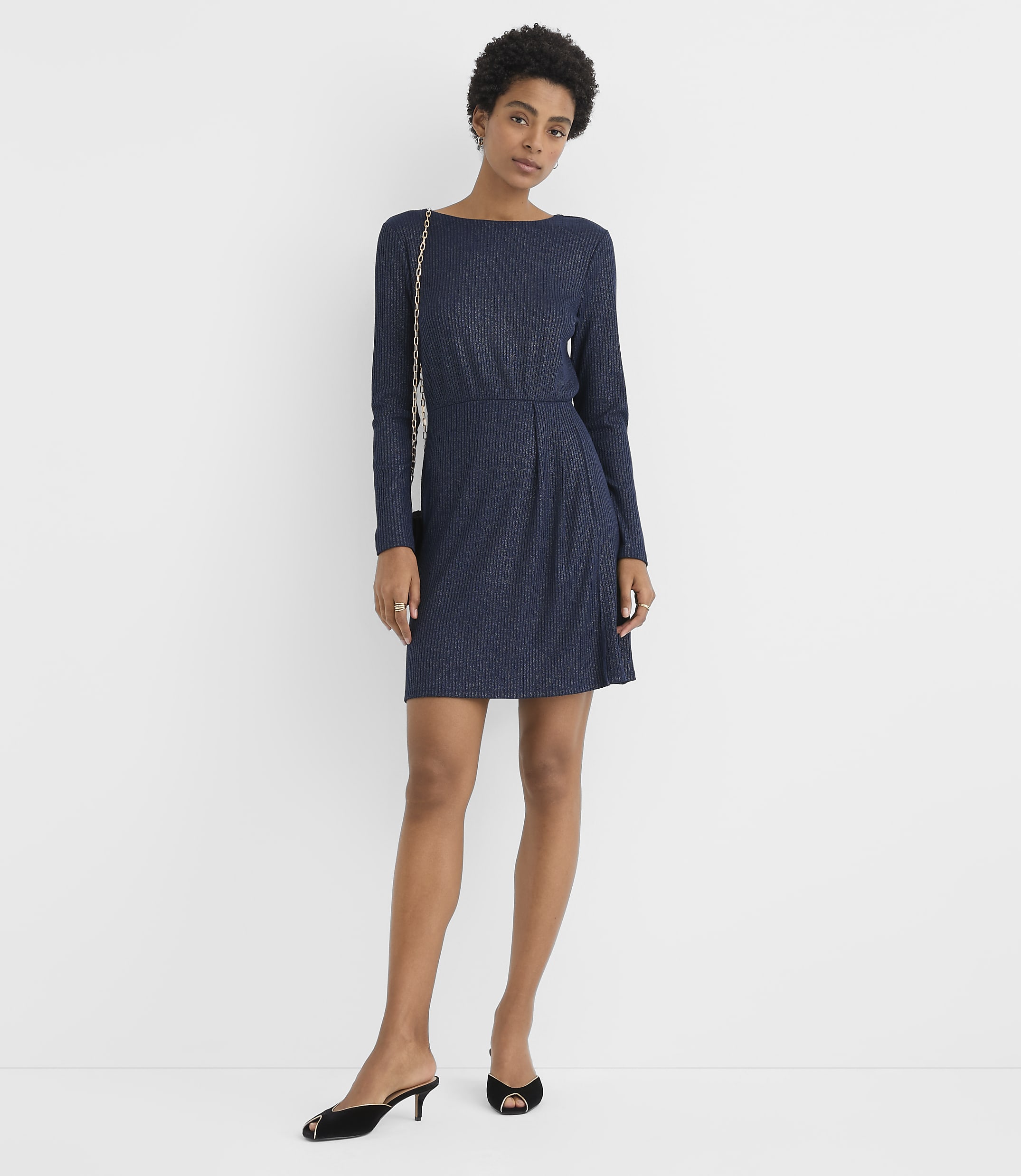 A woman wearing a navy blue long-sleeved dress and black heels stands against a plain white background.