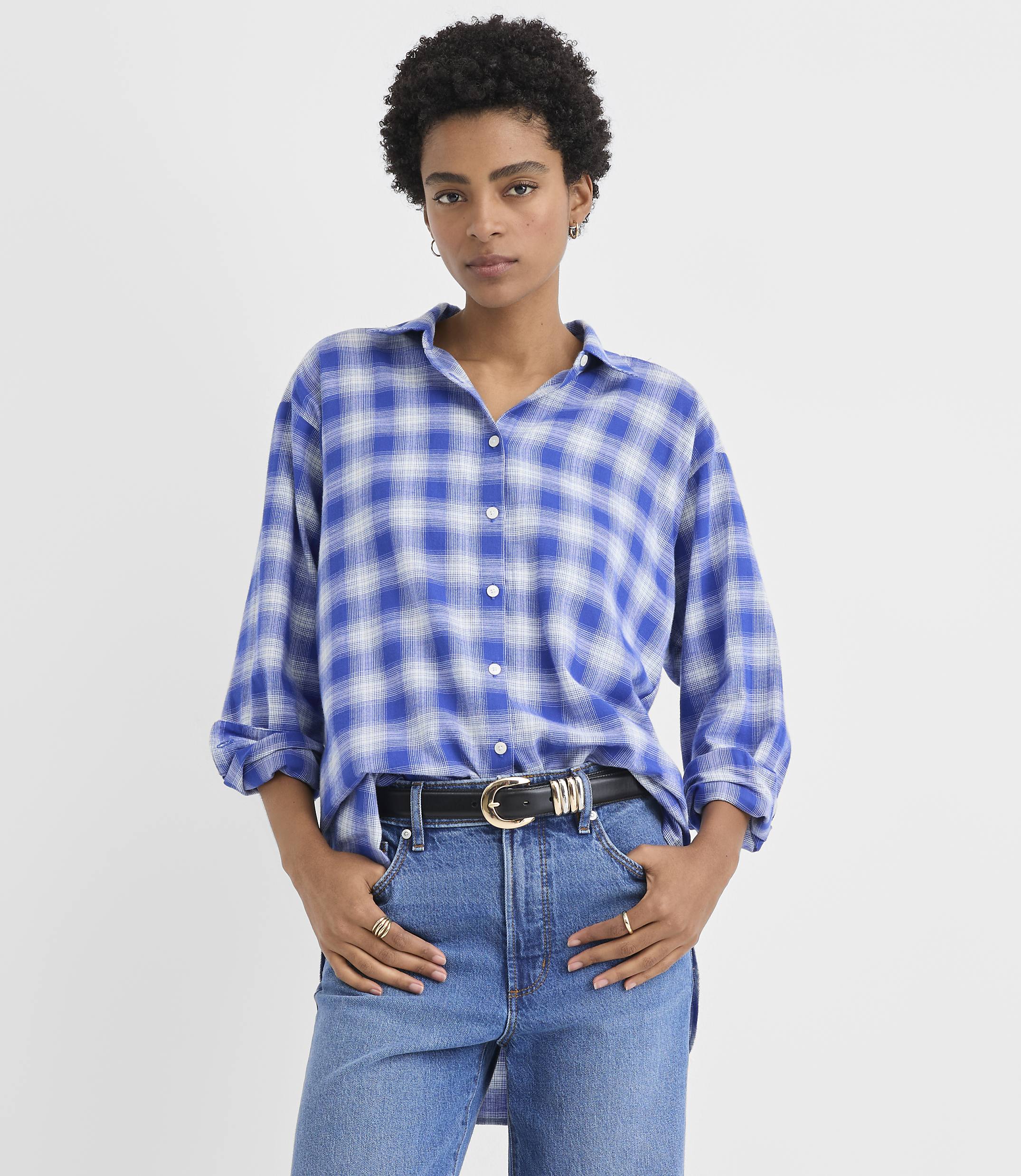 A woman wearing a blue plaid shirt and jeans stands against a plain white background.