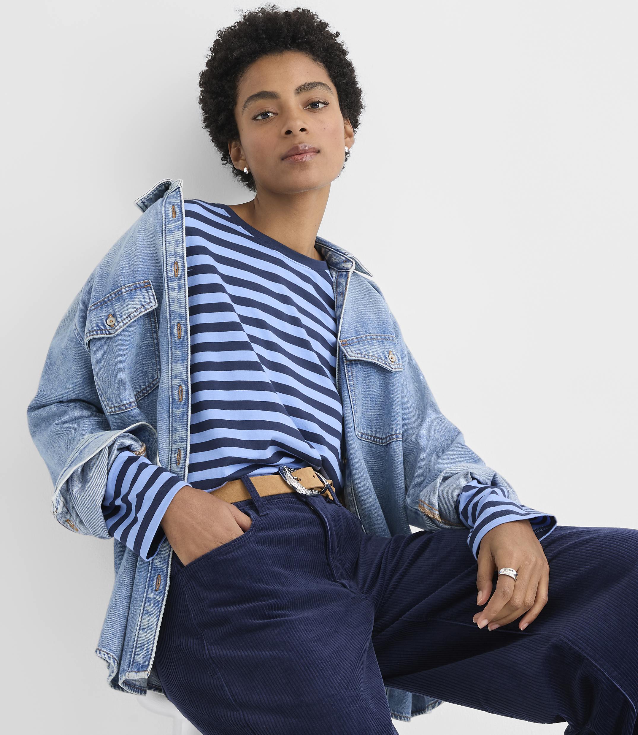 A person wearing a blue striped shirt, a denim jacket, and navy corduroy pants poses against a plain white background.
