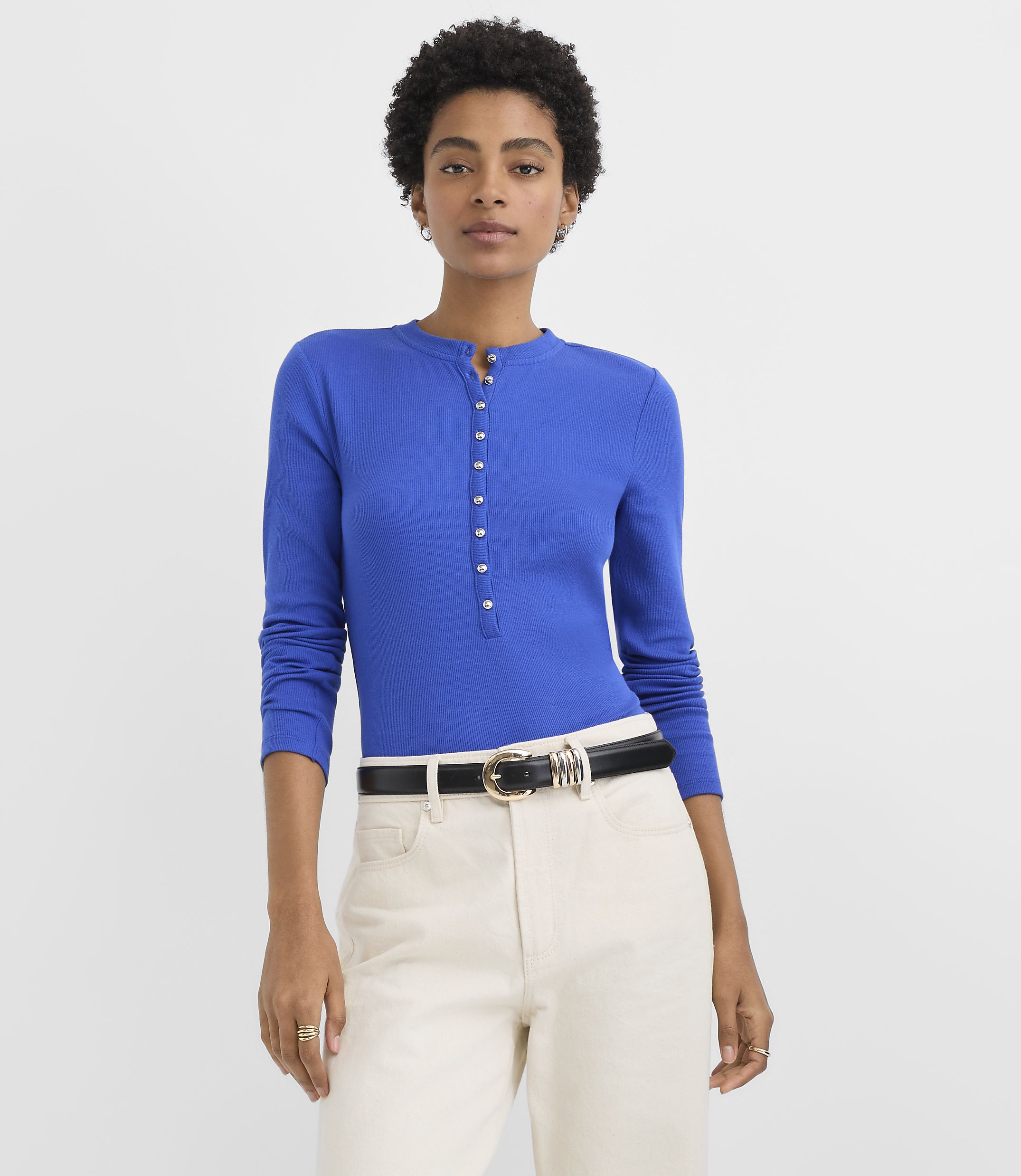A woman wearing a royal blue button-placket top and cream-colored trousers stands against a plain white background.