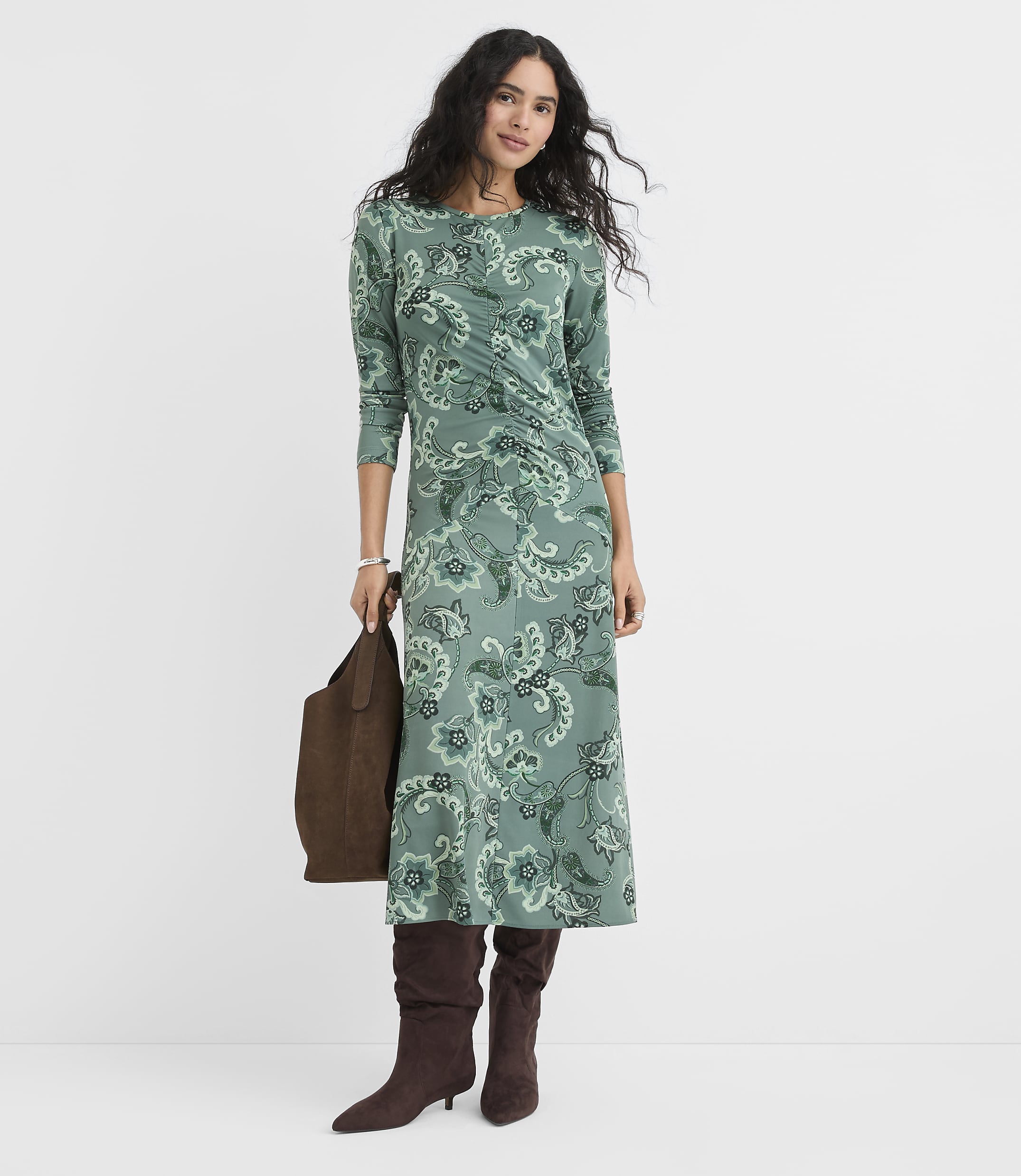 A woman wearing a green floral midi dress and brown boots stands against a plain white background while holding a brown handbag.