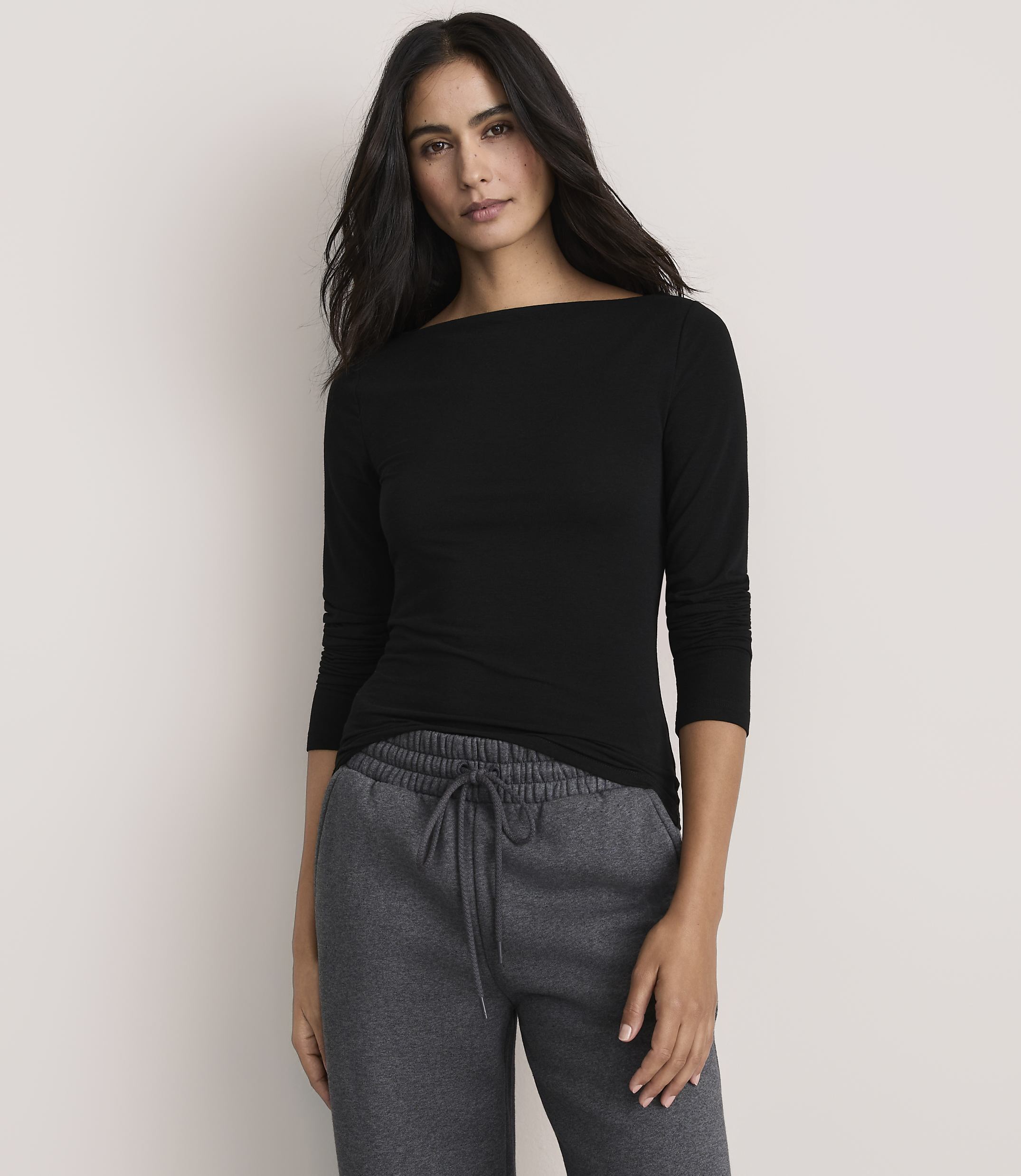A woman wearing a black long-sleeved shirt and grey sweatpants stands against a plain, light-colored background.