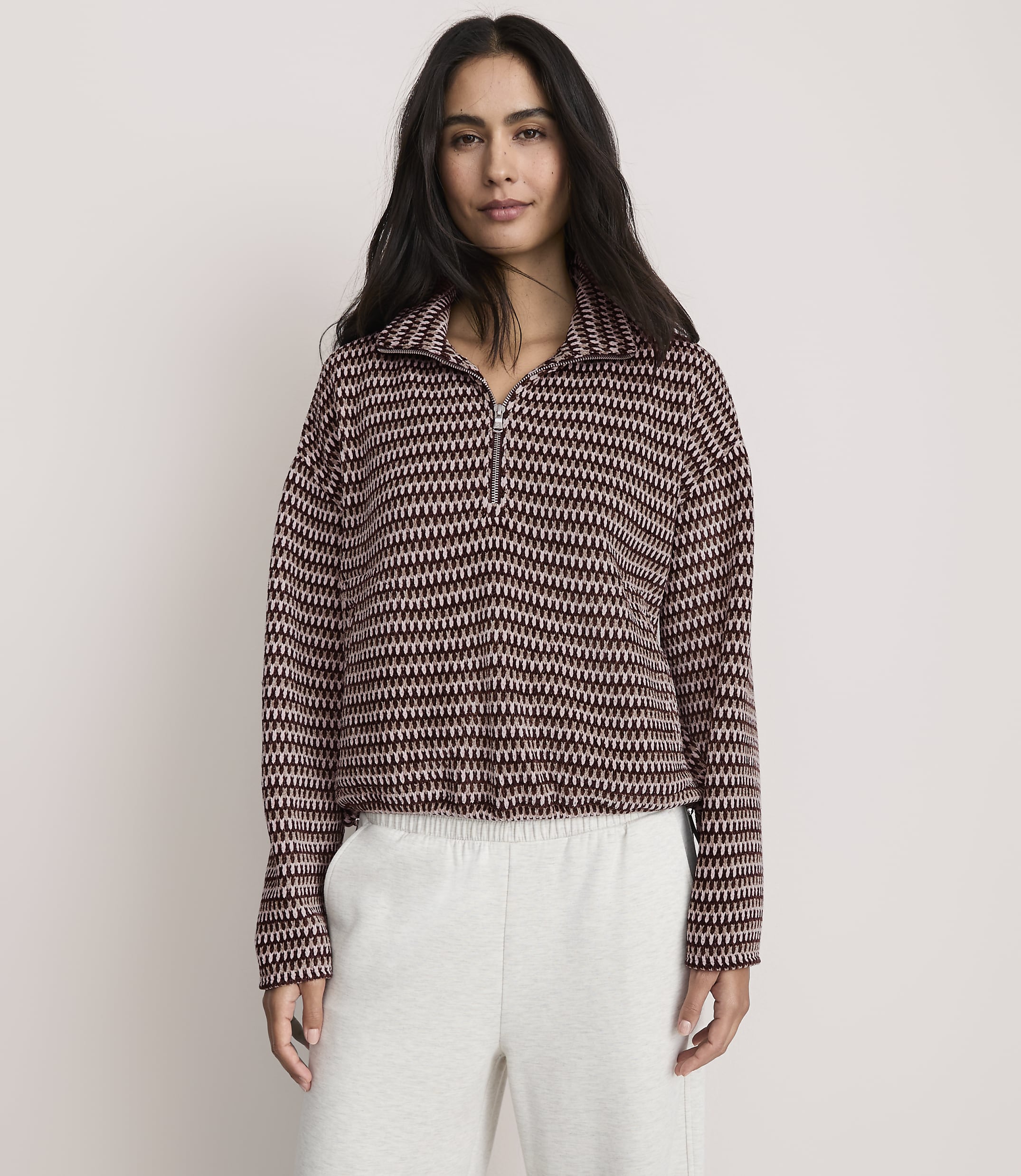 A woman wearing a patterned maroon and white quarter-zip pullover and light grey sweatpants stands against a plain, light-colored background.