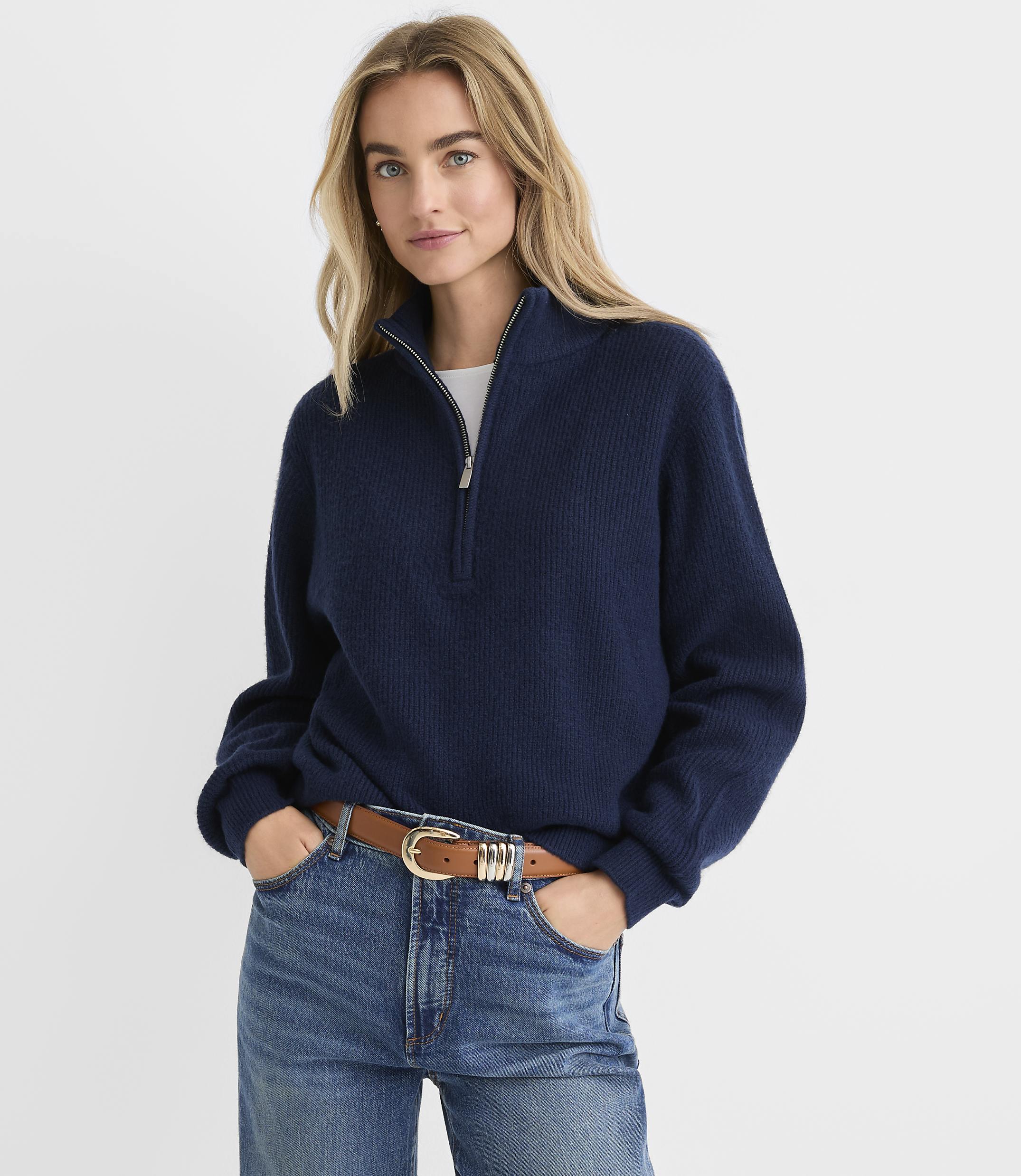 A blonde woman wearing a navy blue quarter-zip sweater and blue jeans stands against a plain white background.