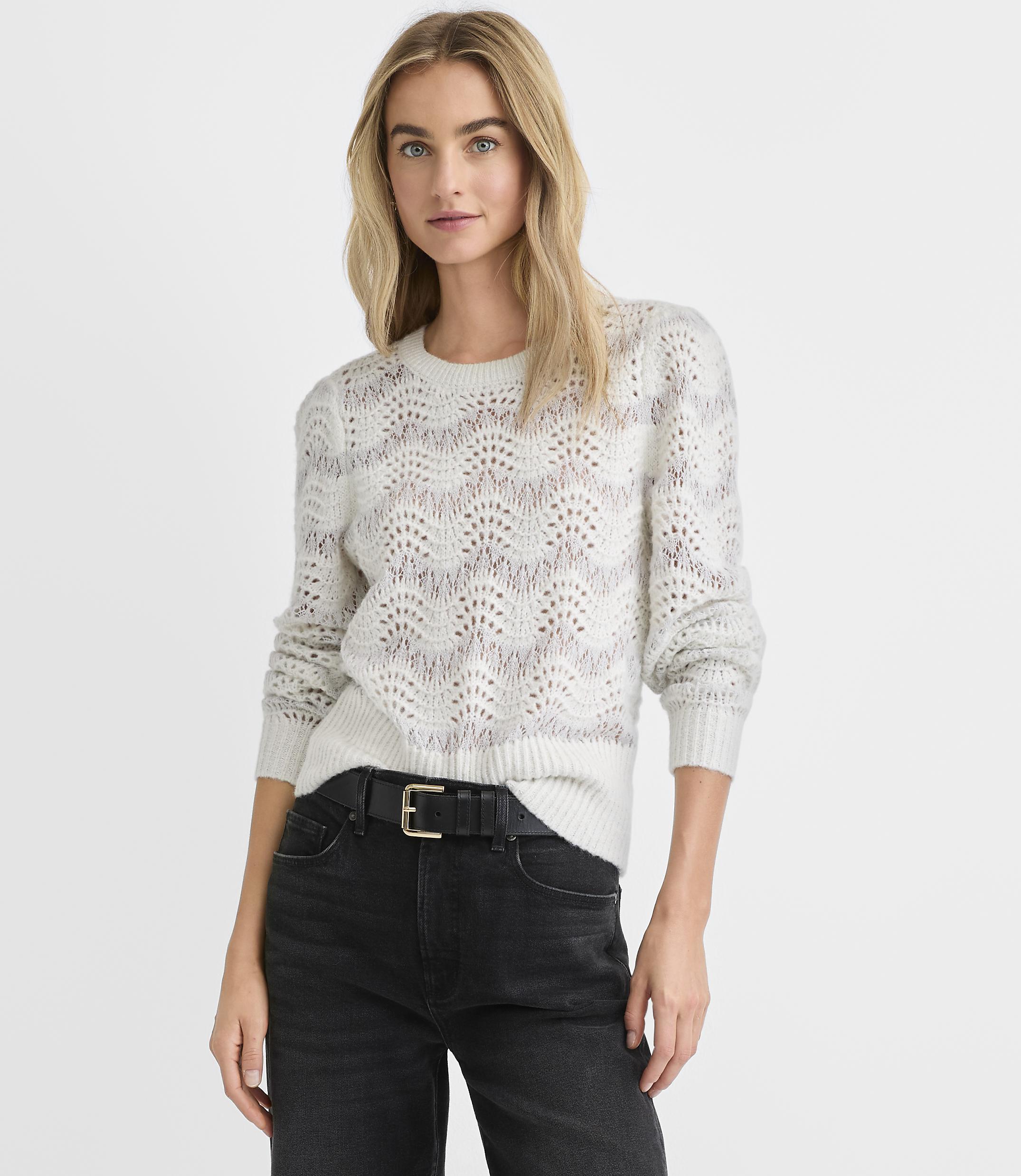 A blonde woman wearing a white pointelle knit sweater and black jeans stands against a plain white background.