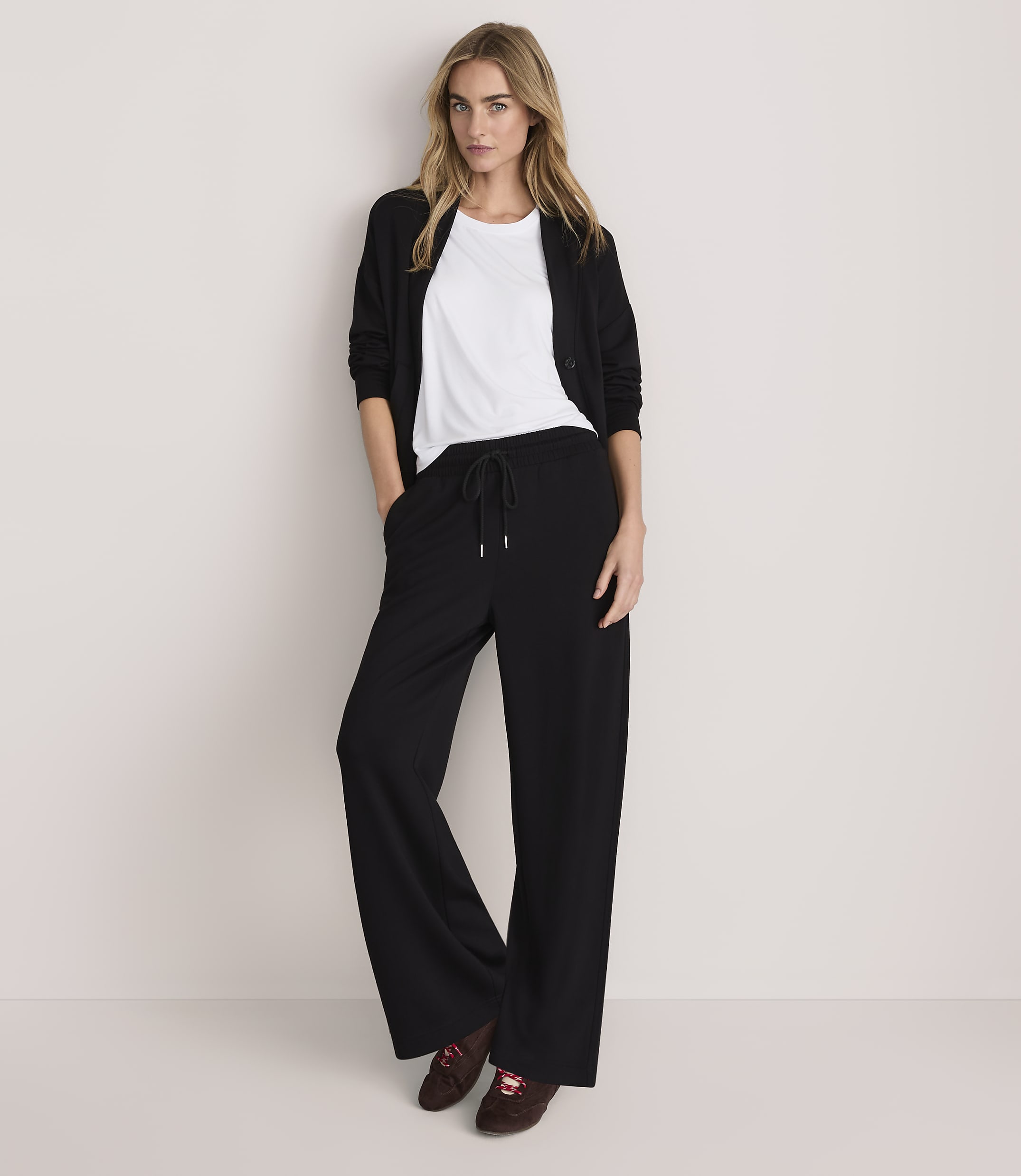 A woman wearing a white t-shirt, black cardigan, and wide-leg black trousers stands against a plain, light-colored background.