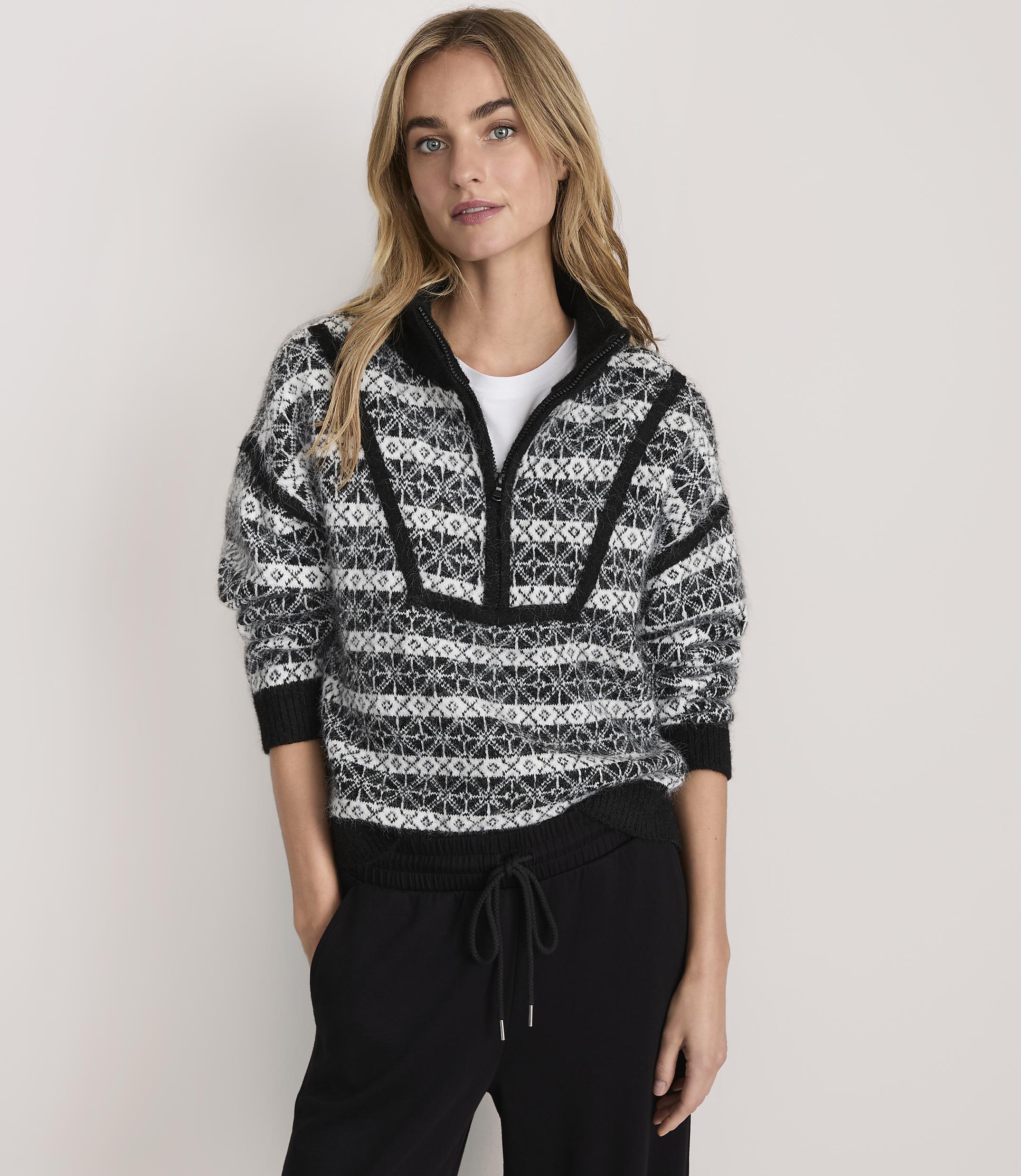 A woman wearing a patterned black and white quarter-zip sweater and black pants stands against a plain, light-colored background.