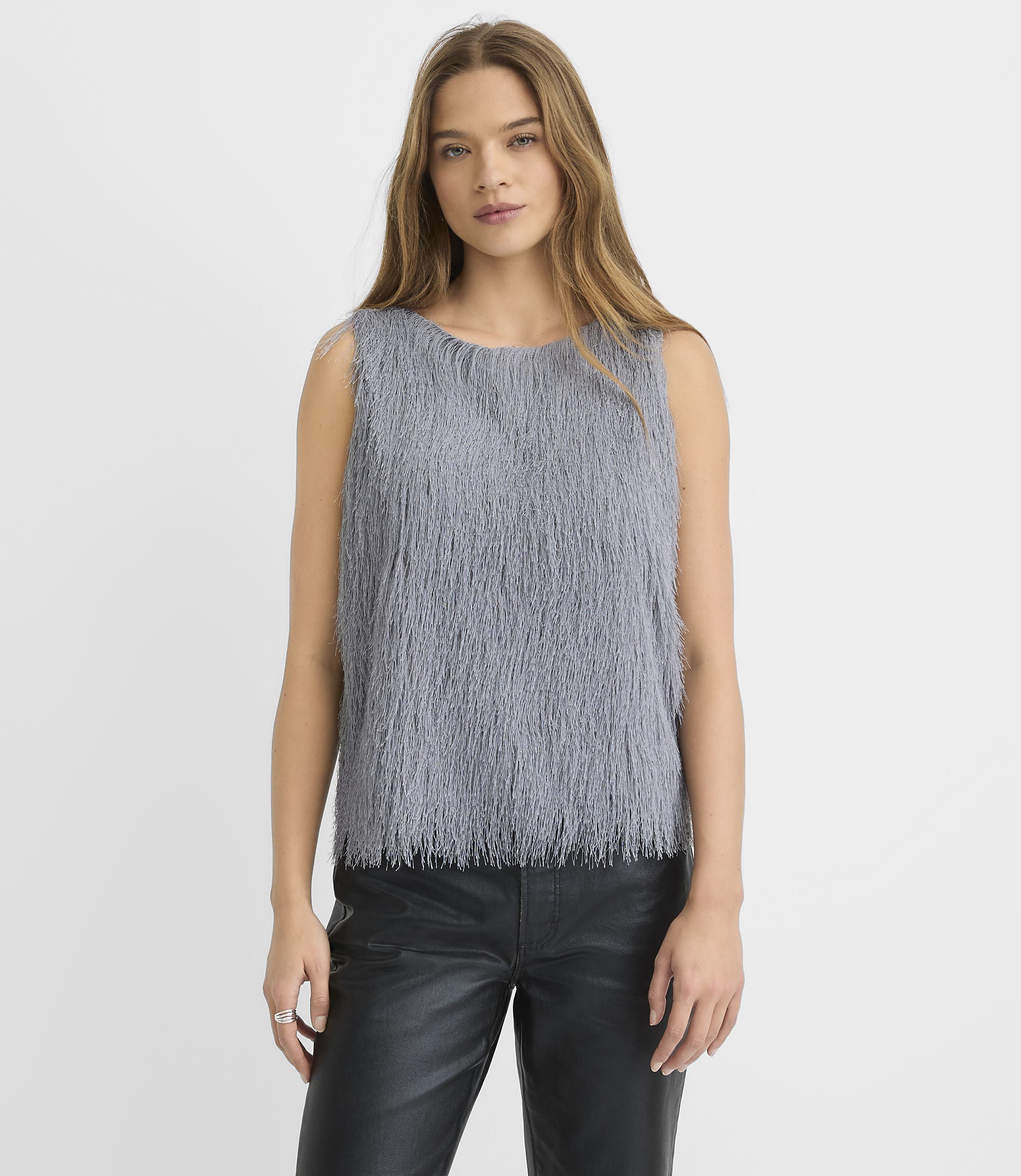 A woman wearing a grey shaggy sleeveless top and black leather pants stands against a plain white background.
