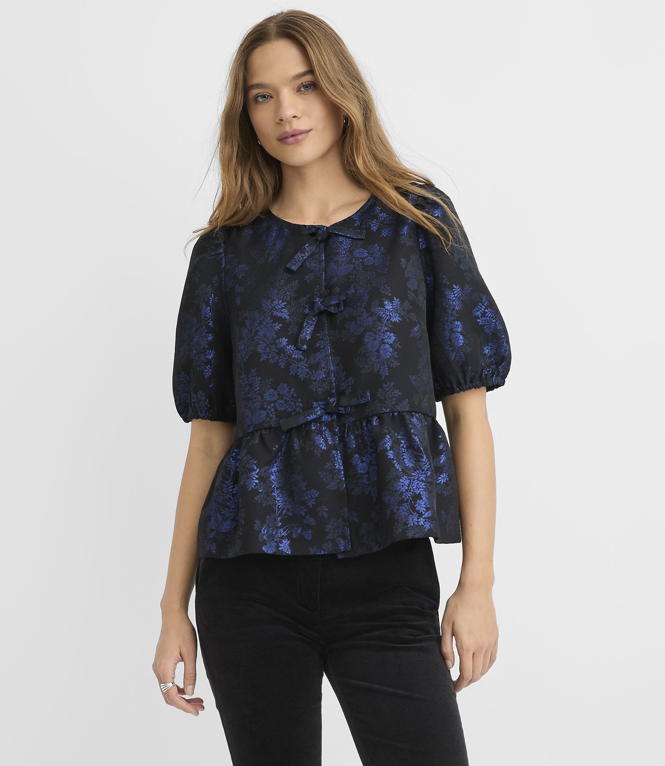 A woman wearing a navy blue floral blouse and black pants stands against a plain light gray background.