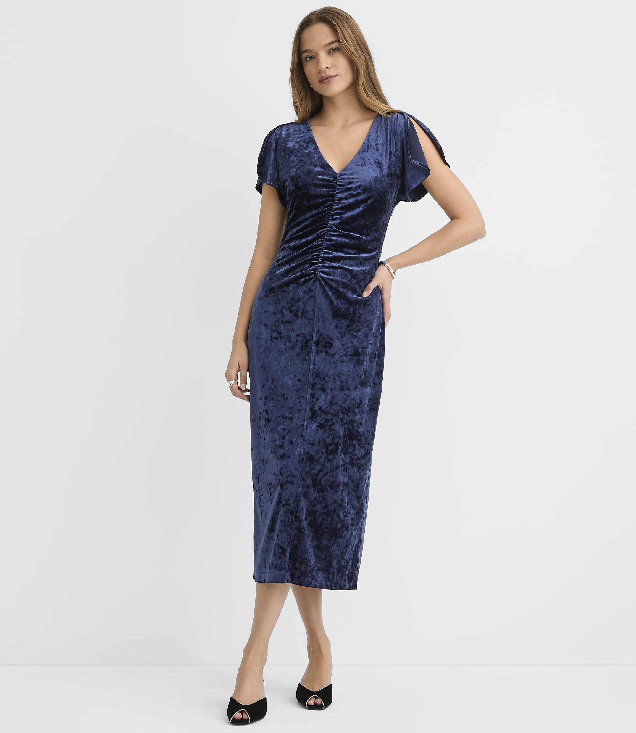 A woman wearing a navy blue velvet midi dress and black heels stands against a plain white background.