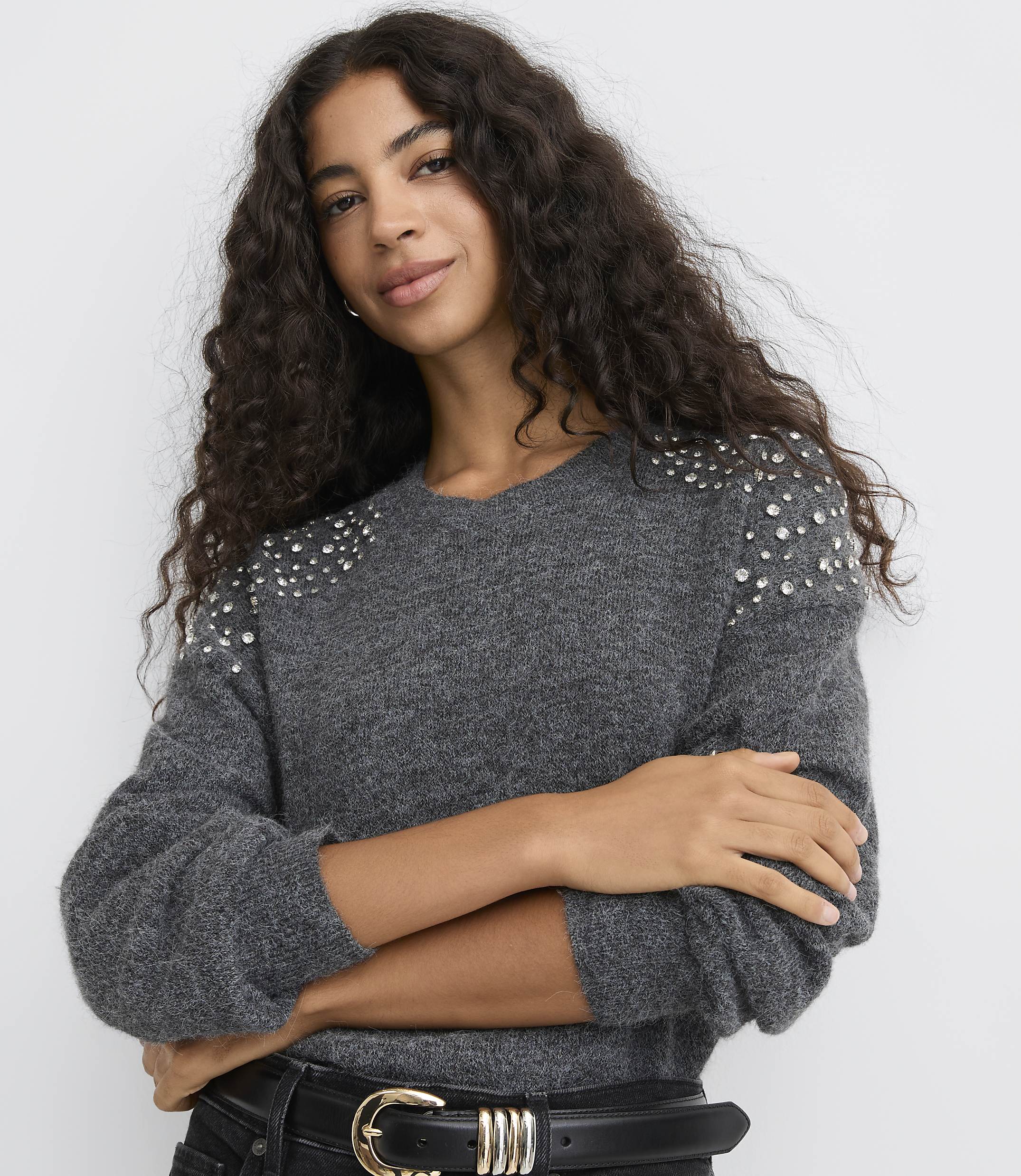 A woman with long curly hair wearing a grey embellished sweater and a black belt poses against a plain white background.