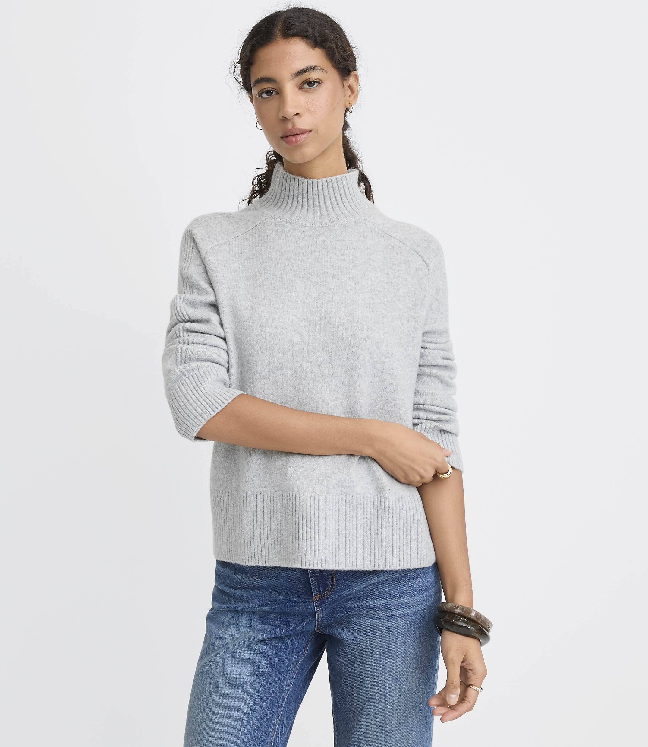 A woman wearing a light grey turtleneck sweater and blue jeans stands against a plain white background.