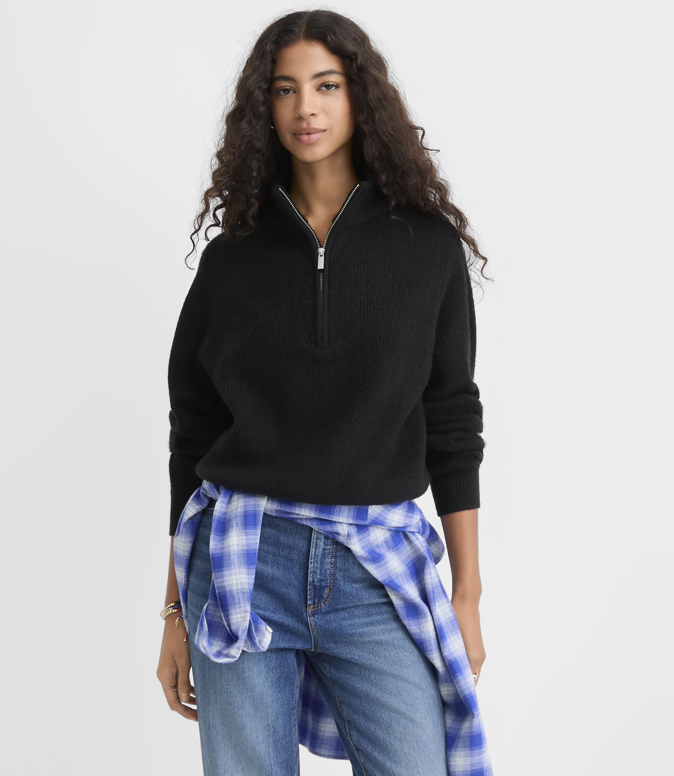 A woman wearing a black quarter-zip sweater and blue jeans with a plaid shirt tied around her waist stands against a plain white background.