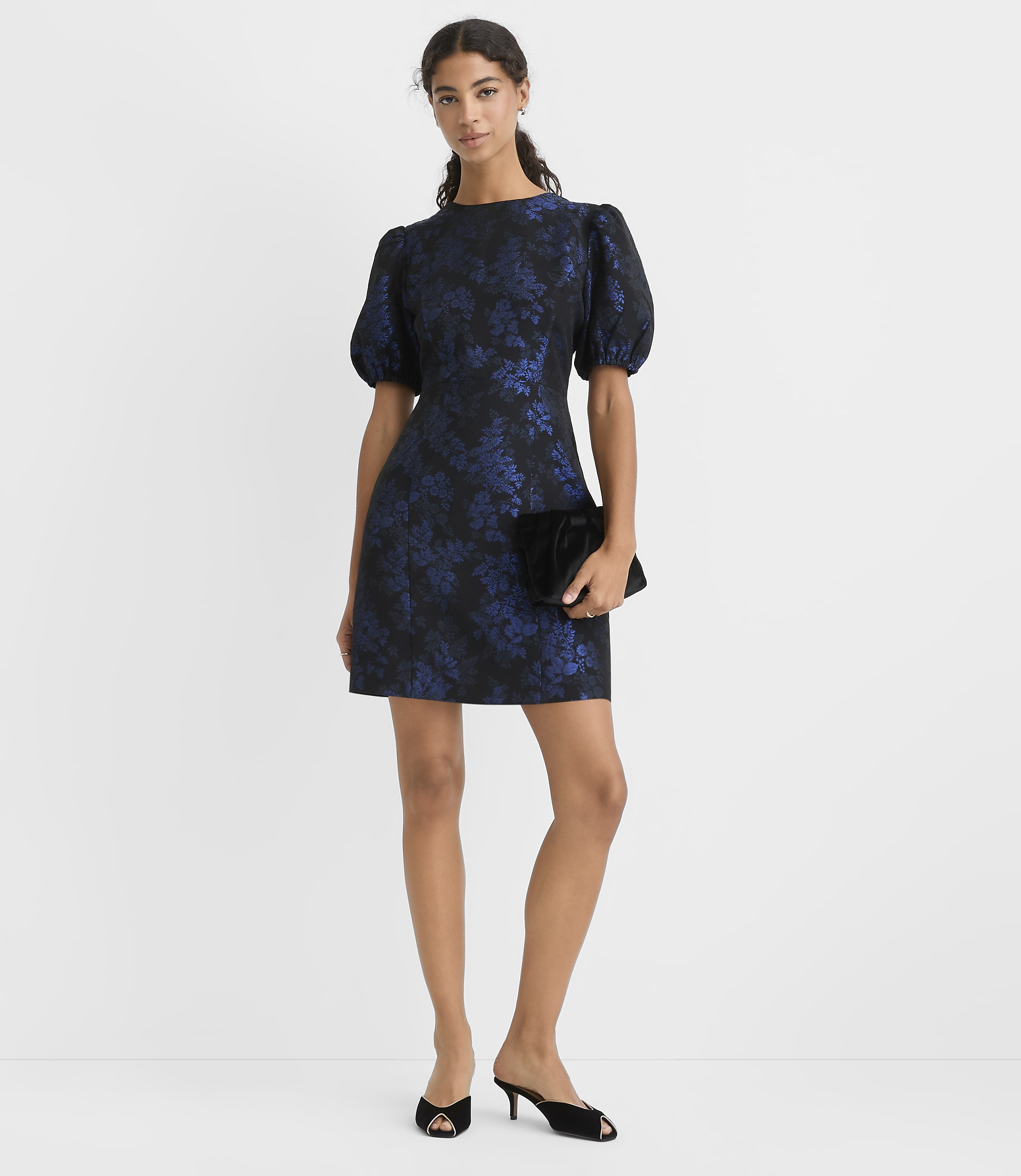 A woman wearing a navy blue floral puff-sleeve dress and black heels stands against a plain white background.