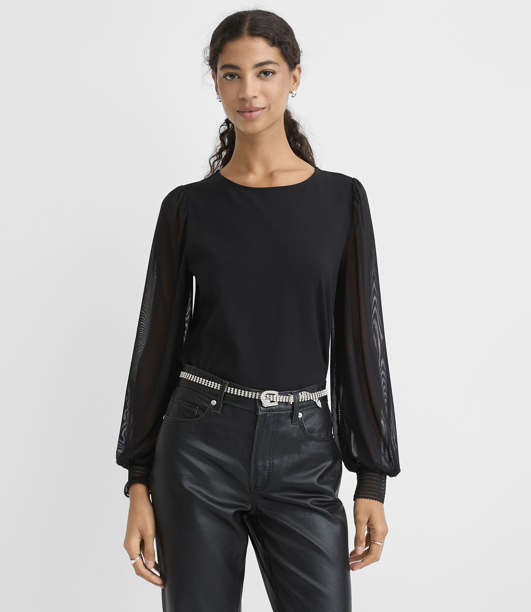 A woman wearing a black top with sheer sleeves and black faux-leather pants stands against a plain white background.