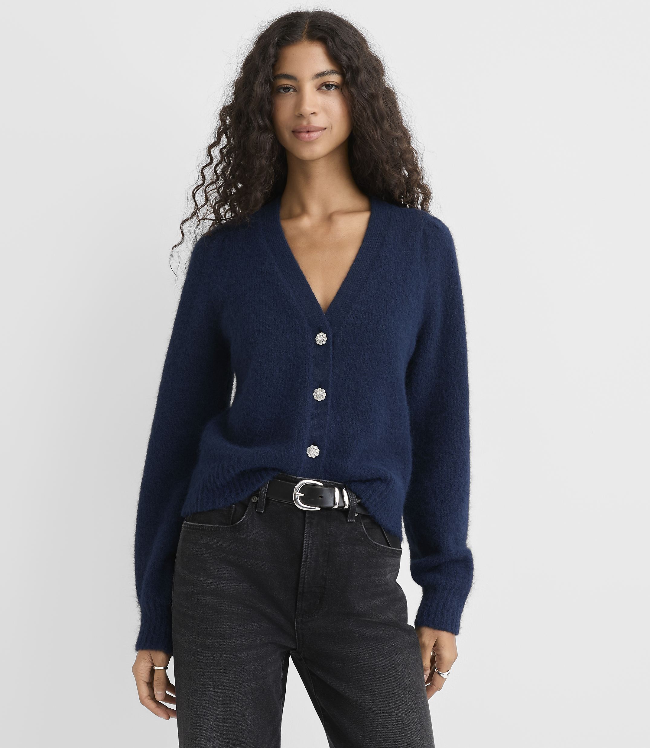 A woman wearing a navy blue cardigan and black jeans stands against a plain light gray background.