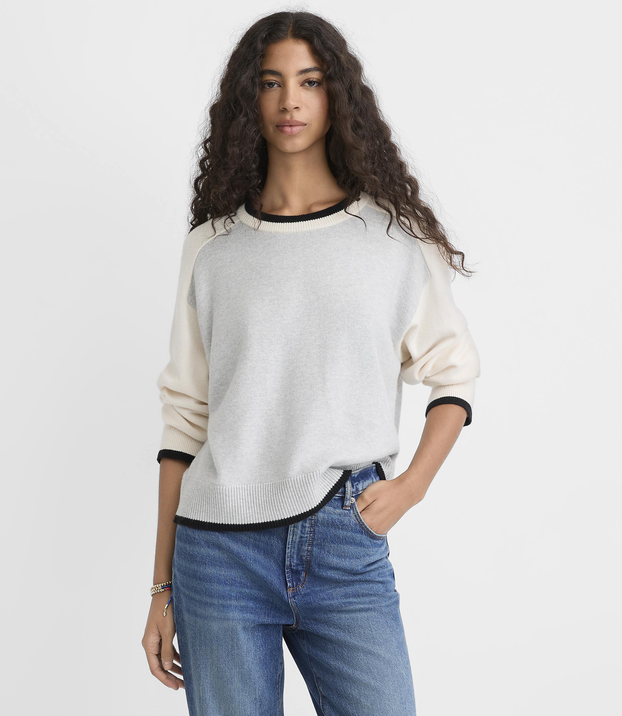 A woman with long curly hair wearing a two-tone sweater and blue jeans stands against a plain white background.