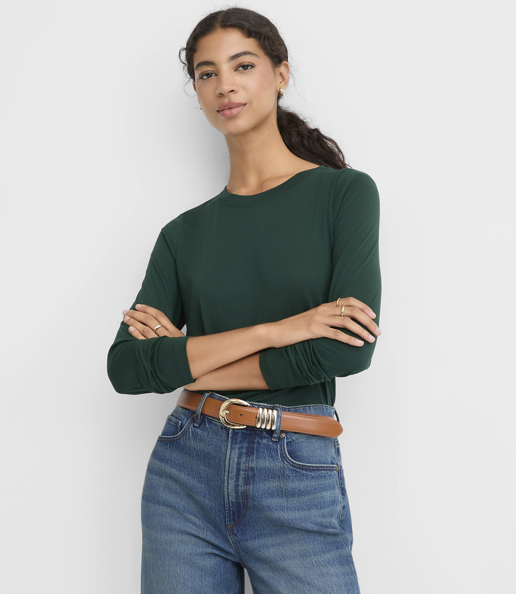 A woman wearing a dark green long-sleeved shirt and blue jeans with a brown belt stands with her arms crossed against a plain white background.