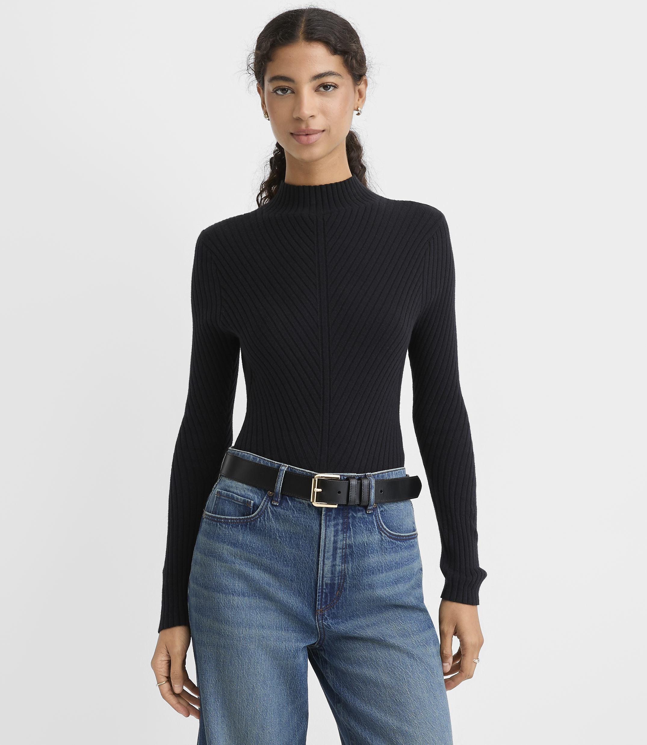A woman wearing a black ribbed mock-neck sweater, blue jeans, and a black belt stands against a plain light gray background.