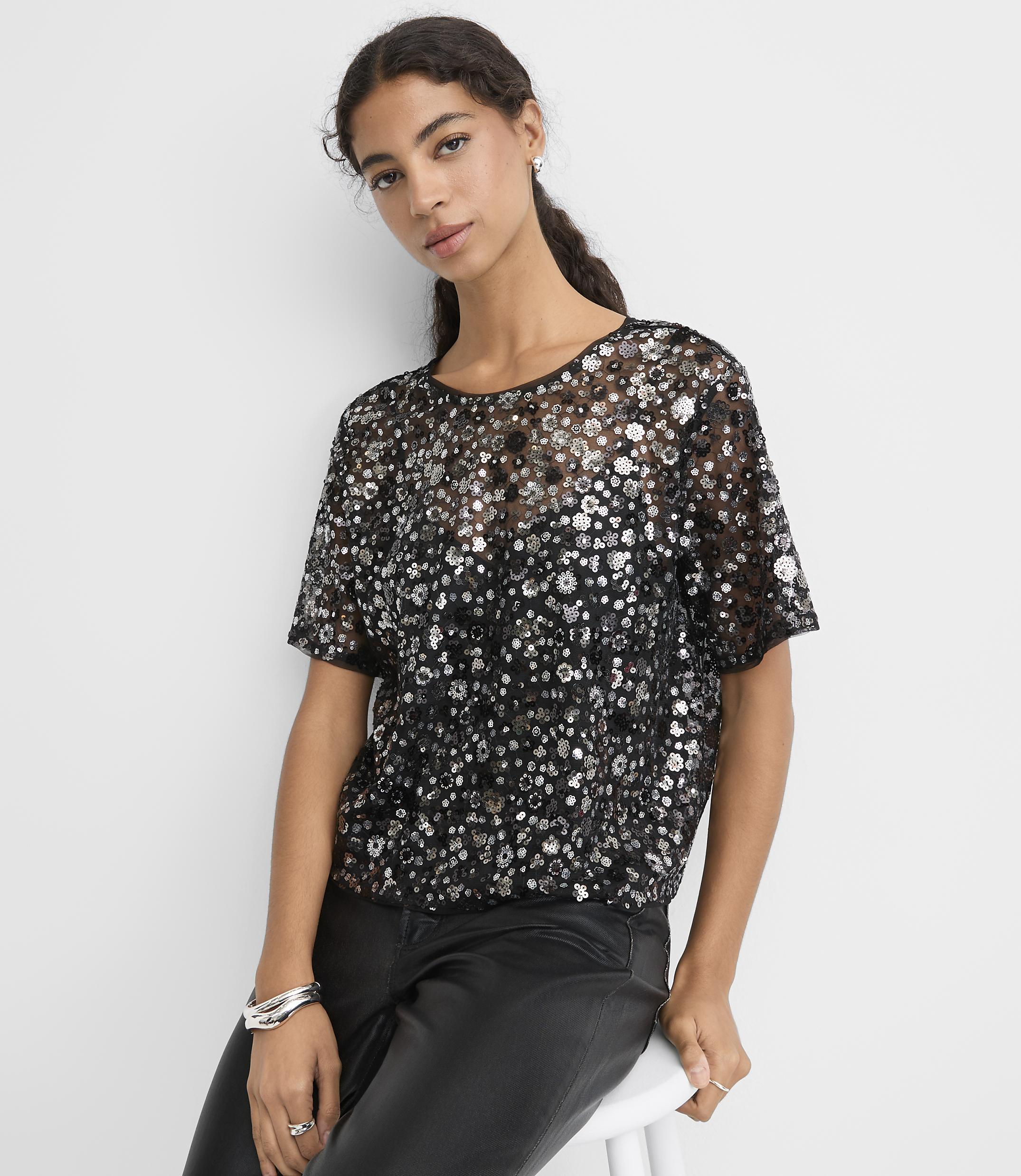 A woman wearing a sequined black top and leather pants sits against a plain white background.