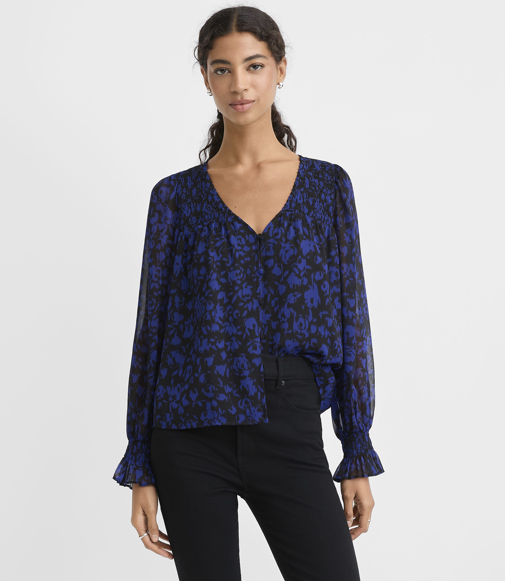 A woman wearing a blue and black floral print blouse and black pants stands against a plain white background.
