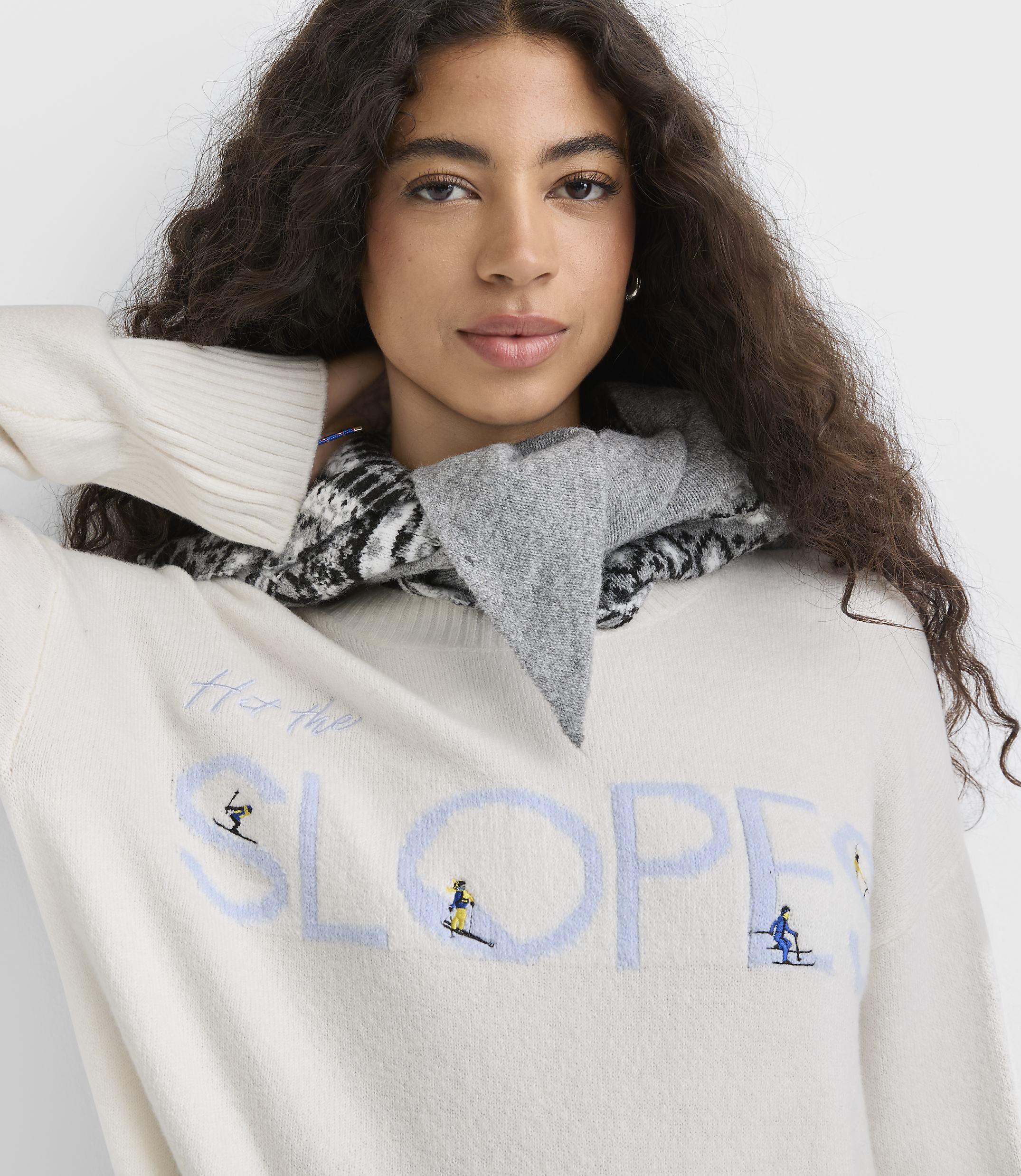 A woman with long curly hair wearing a white graphic sweater and a grey patterned scarf poses against a plain white background.