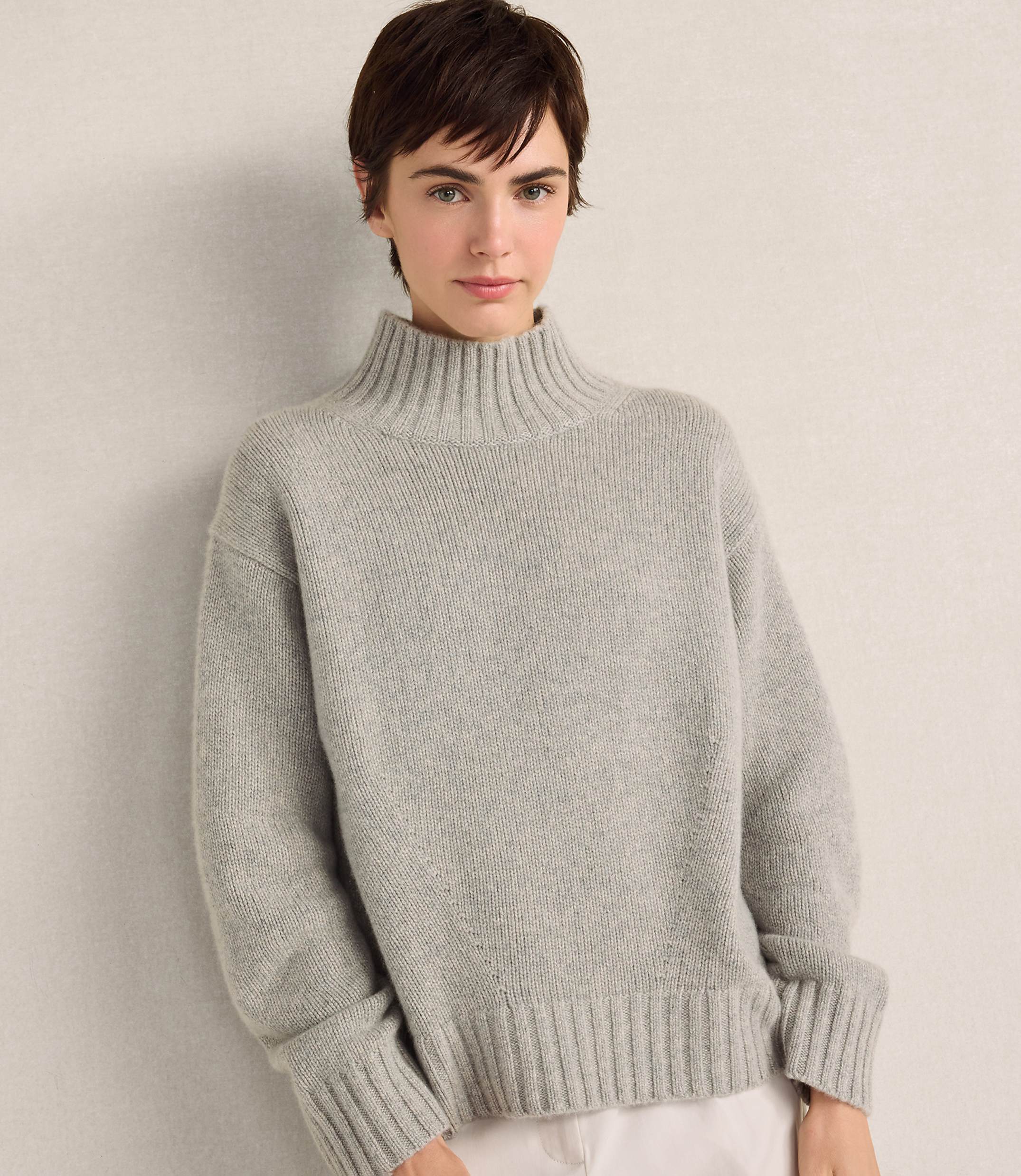 A woman with short brown hair wearing a grey ribbed turtleneck sweater stands against a plain, light-colored wall.