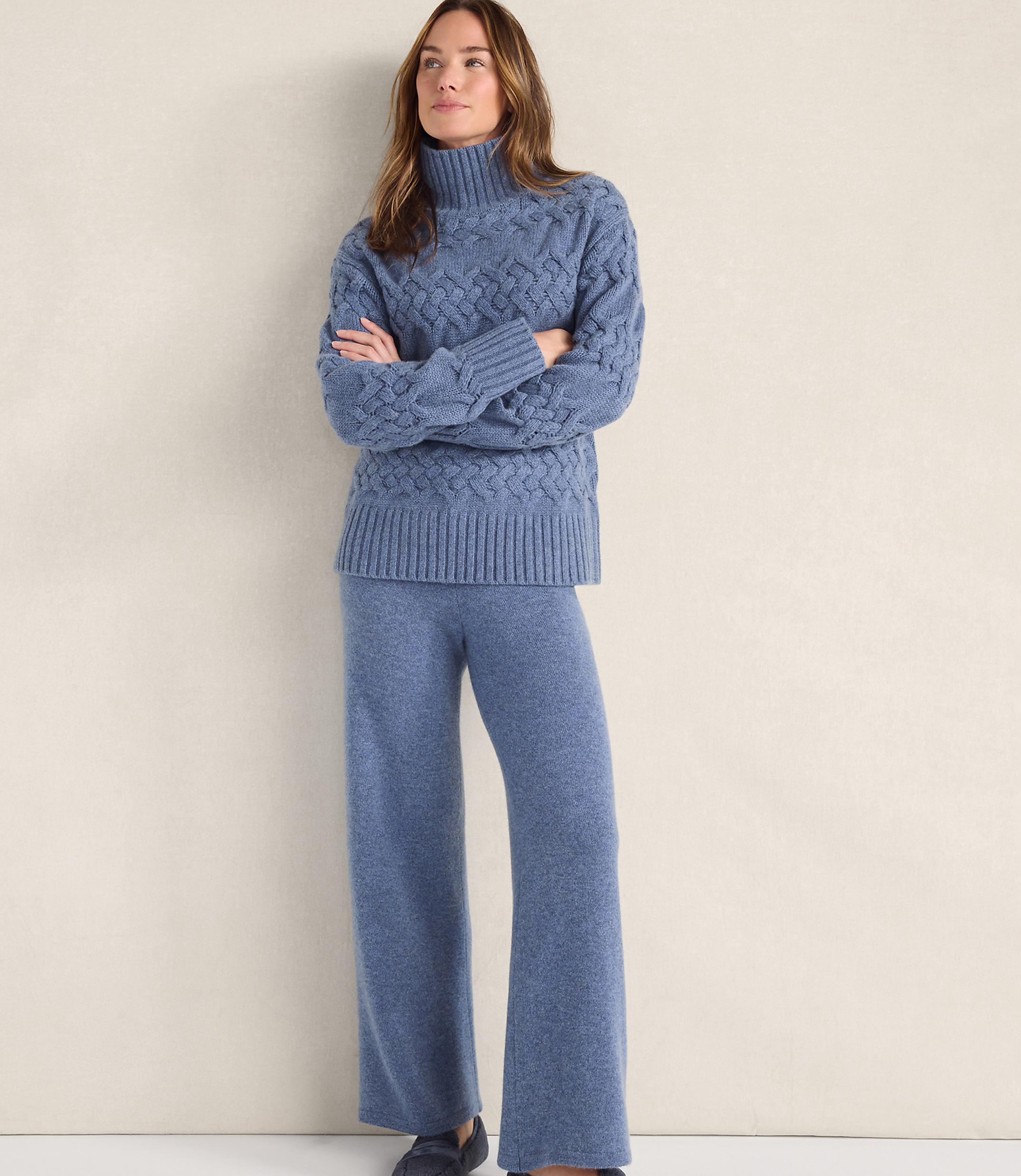 A woman wearing a blue cable-knit sweater and matching wide-leg trousers stands against a plain, light-colored background.