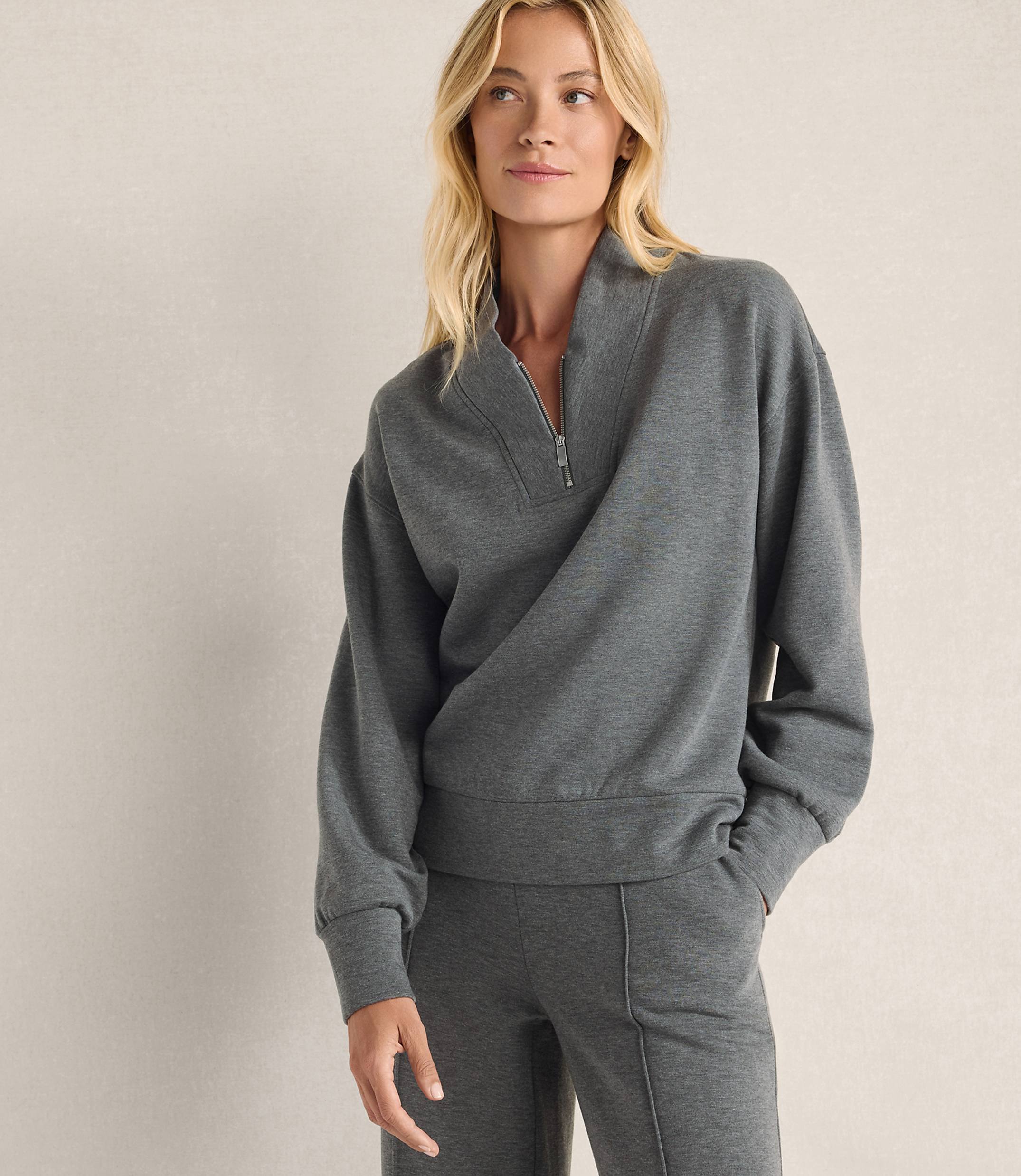 A blonde woman wearing a grey quarter-zip sweatshirt and matching pants stands against a plain, light-colored wall.