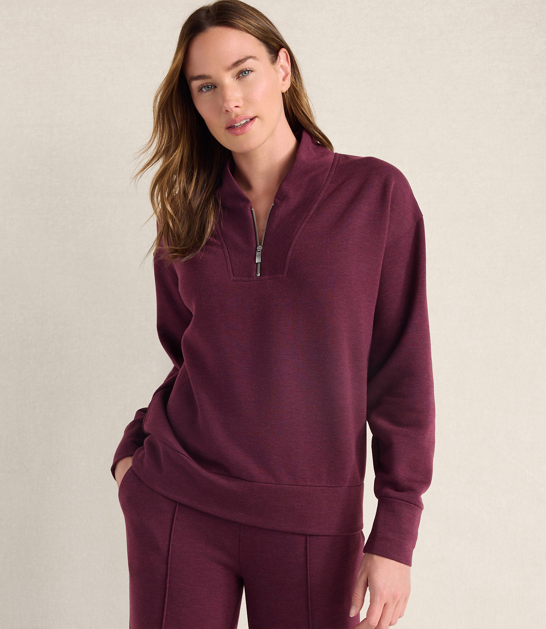 A woman wearing a maroon quarter-zip pullover and matching pants stands against a plain, light-colored wall.