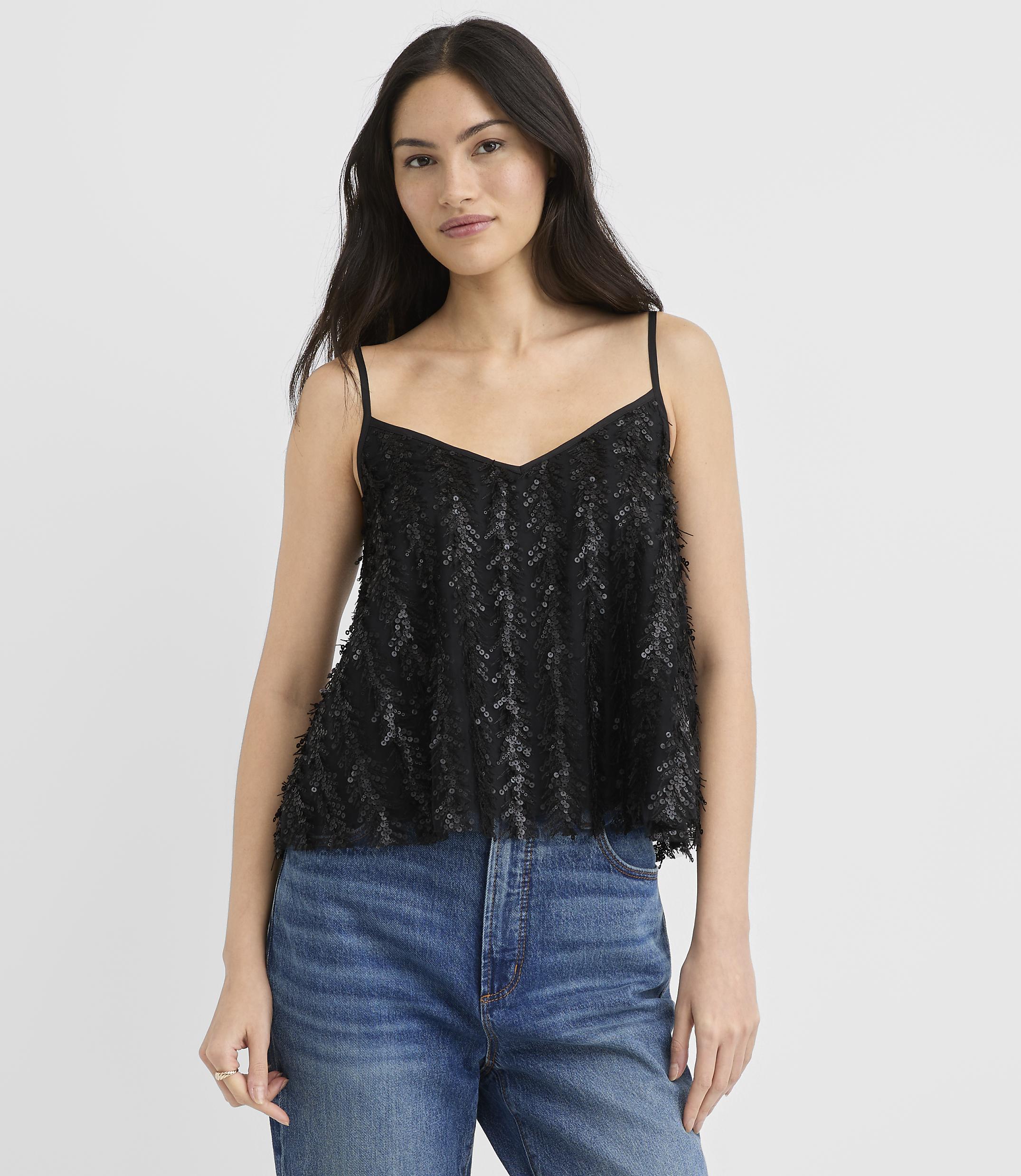 A woman wearing a black sequined camisole and blue jeans stands against a plain white background.