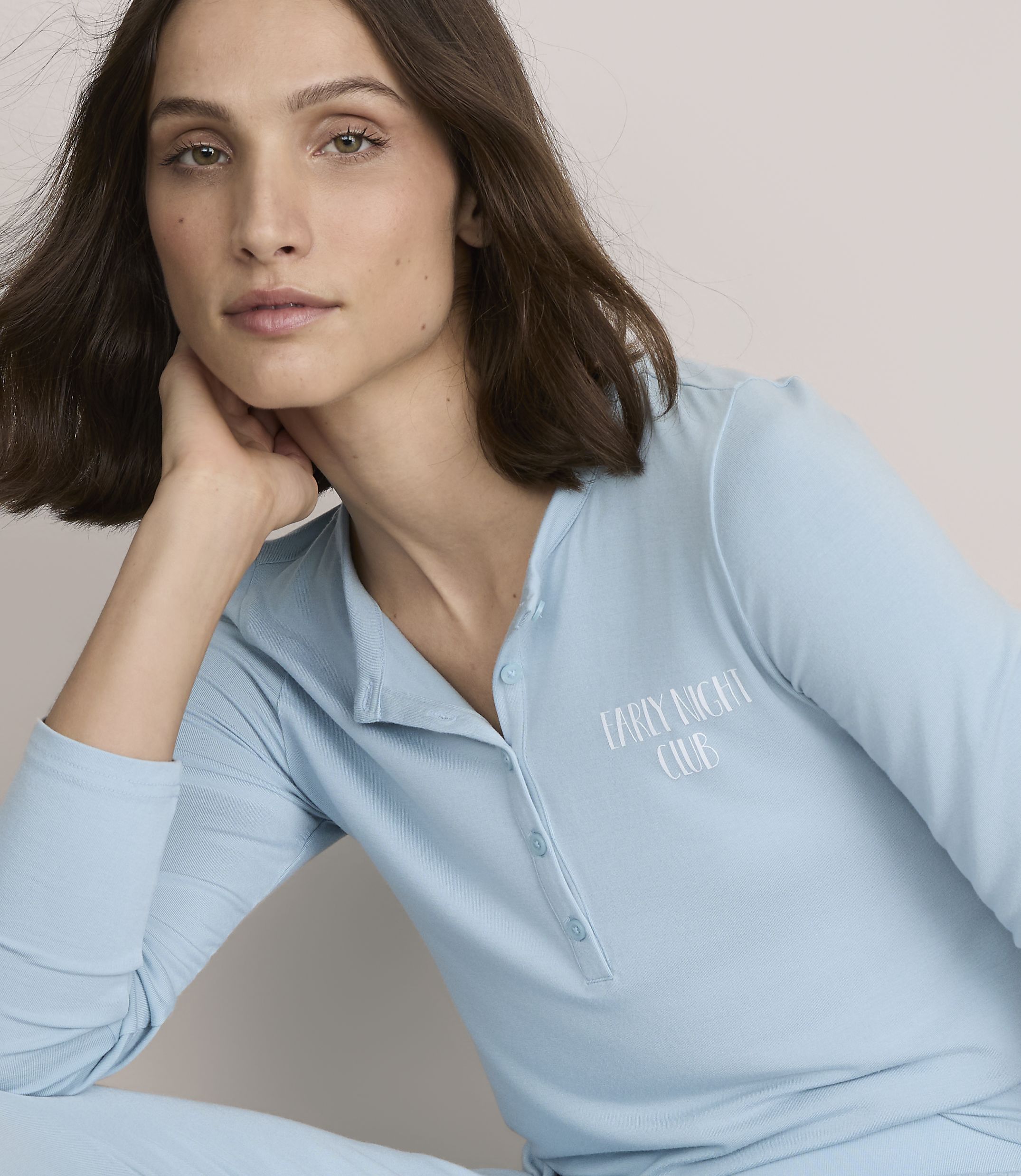 A woman with brown hair wearing light blue pajamas rests her chin on her hand against a plain, neutral background.