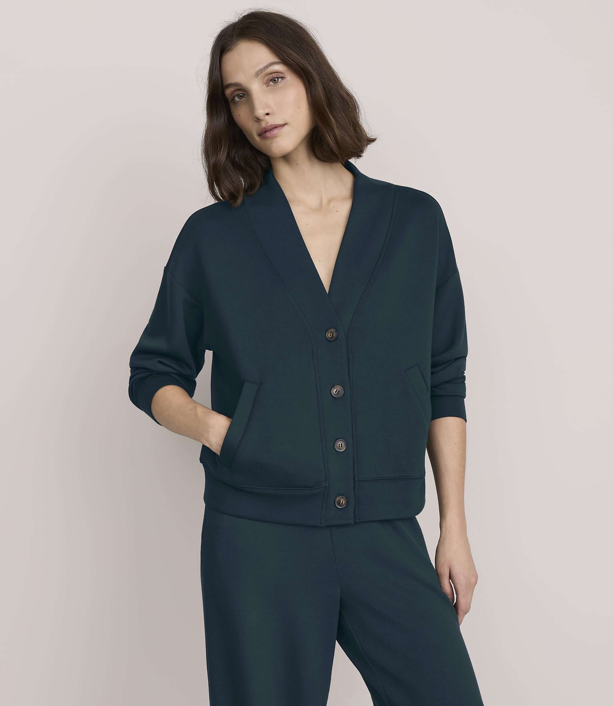 A woman wearing a dark teal button-up cardigan and matching trousers stands against a plain, light beige background.