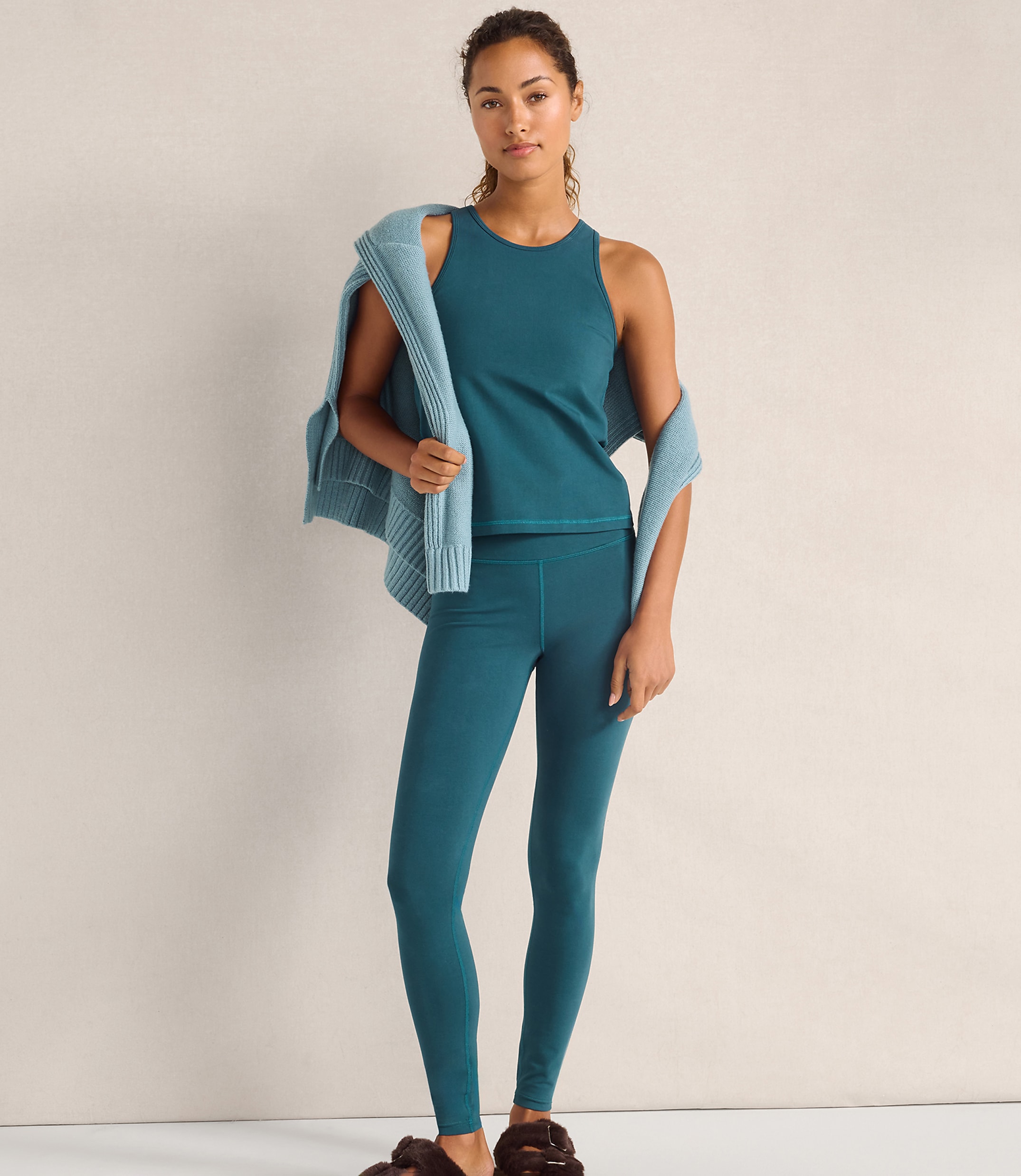 A woman wearing a teal tank top and leggings with a light blue cardigan draped over her shoulders stands against a plain, neutral-colored wall.