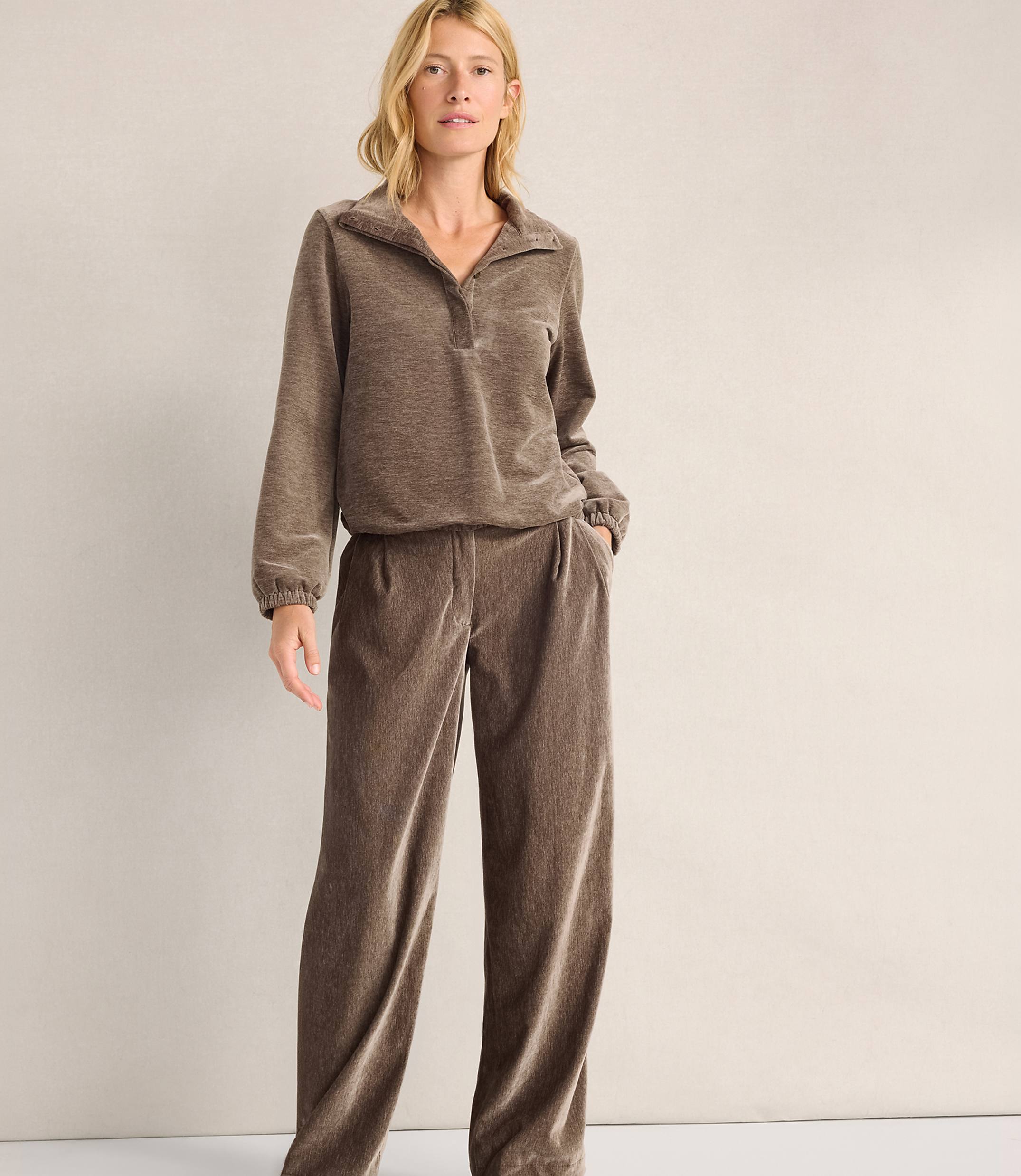A woman wearing a brown velvet lounge set stands against a plain, light-colored wall.