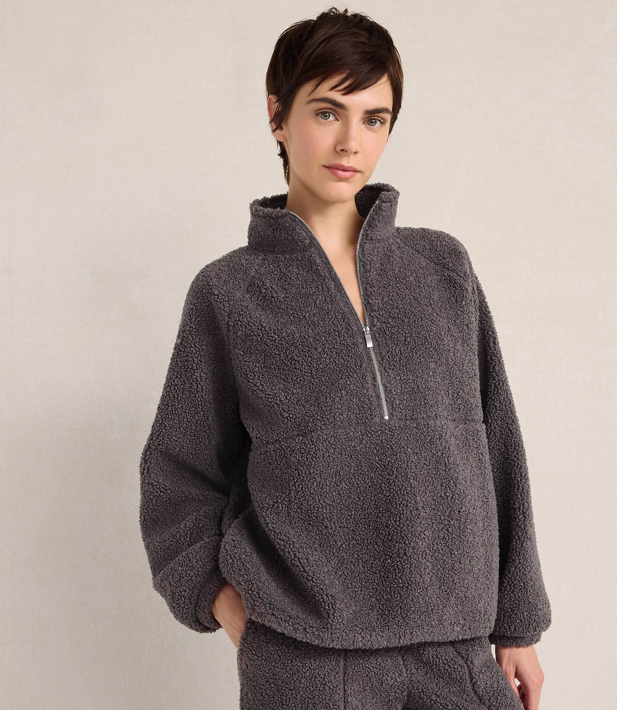 A woman with short brown hair wears a charcoal grey fleece quarter-zip pullover and matching pants against a plain, light-colored background.