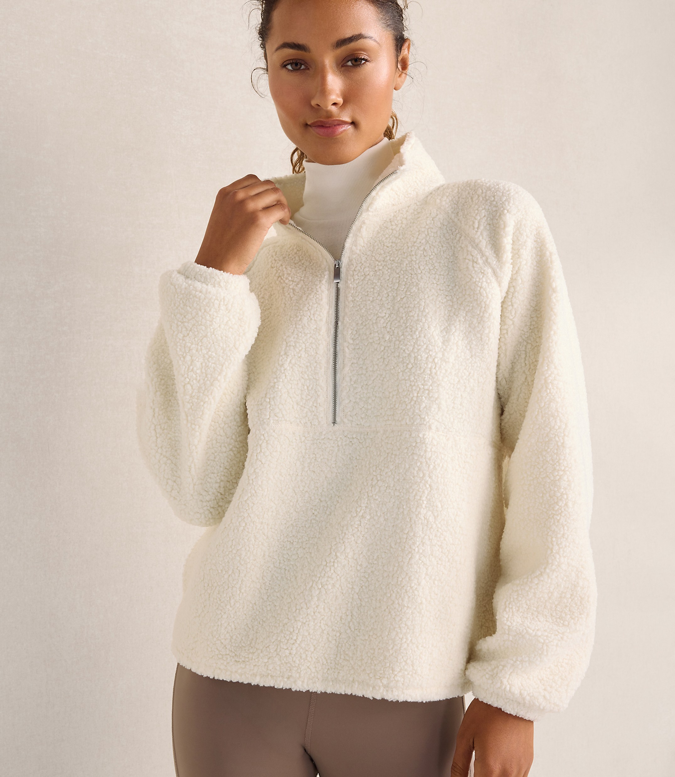 A woman wearing a white fleece pullover and a white turtleneck stands against a plain, neutral-toned background.