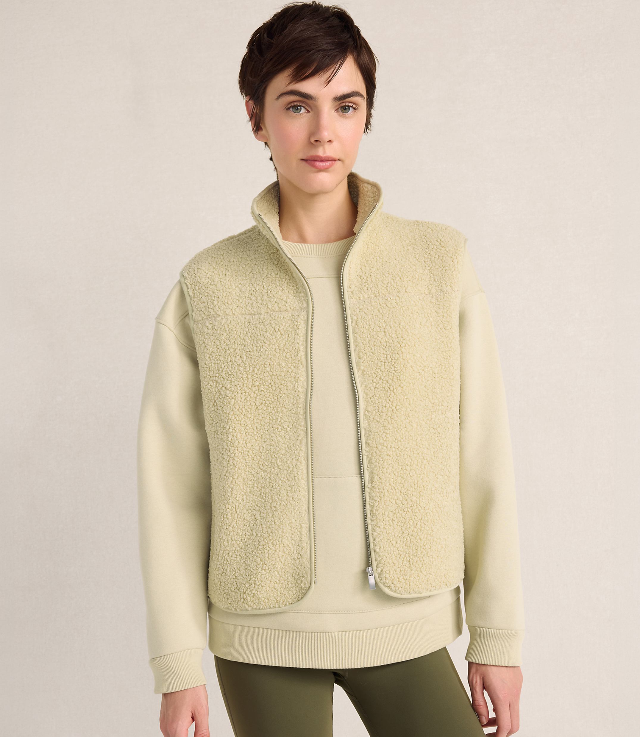 A woman with short dark hair wears a cream-colored fleece vest over a matching sweatshirt and olive green leggings against a plain, neutral background.