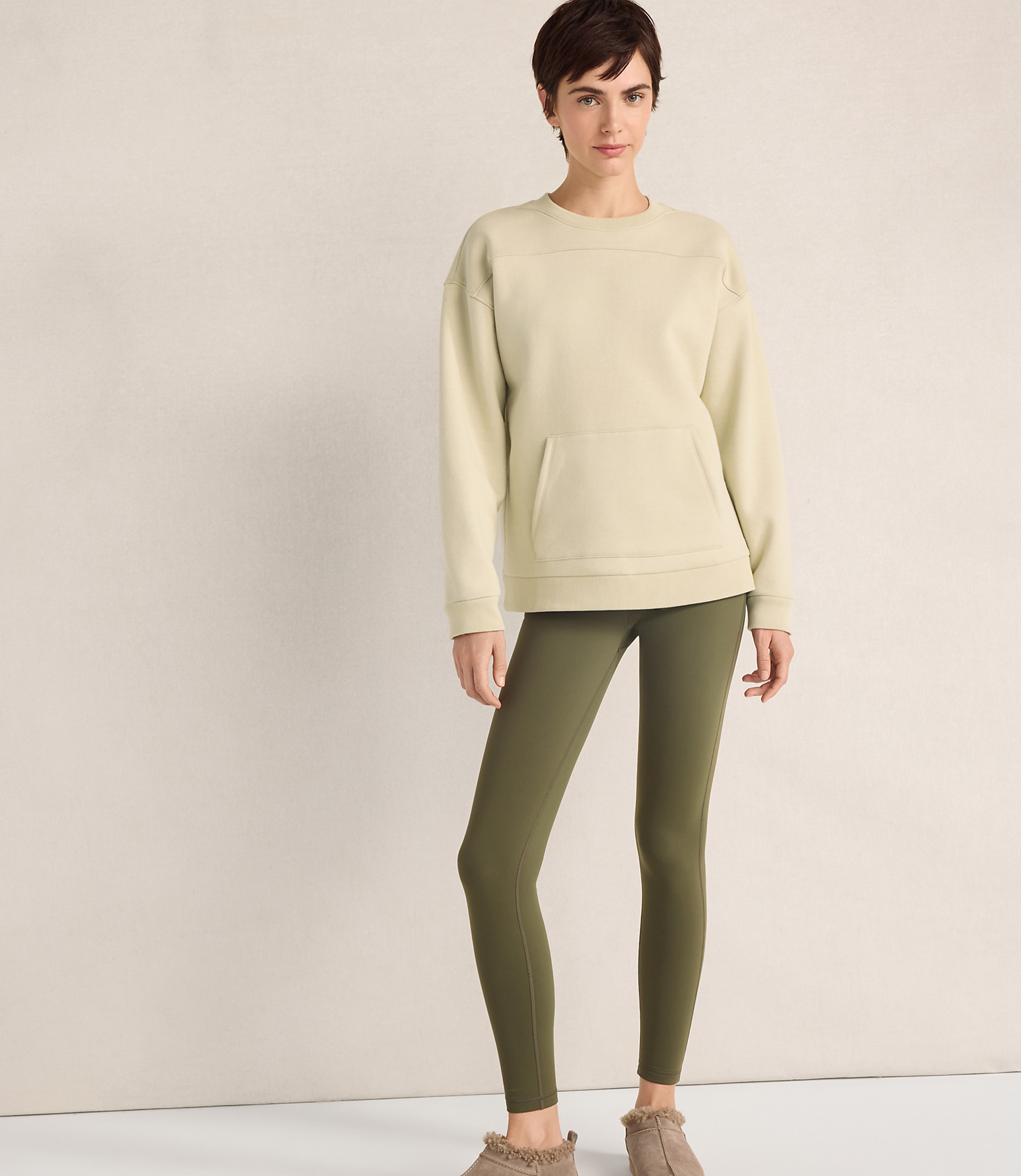 A woman wearing a cream-colored sweatshirt and olive green leggings stands against a plain, neutral-toned wall.