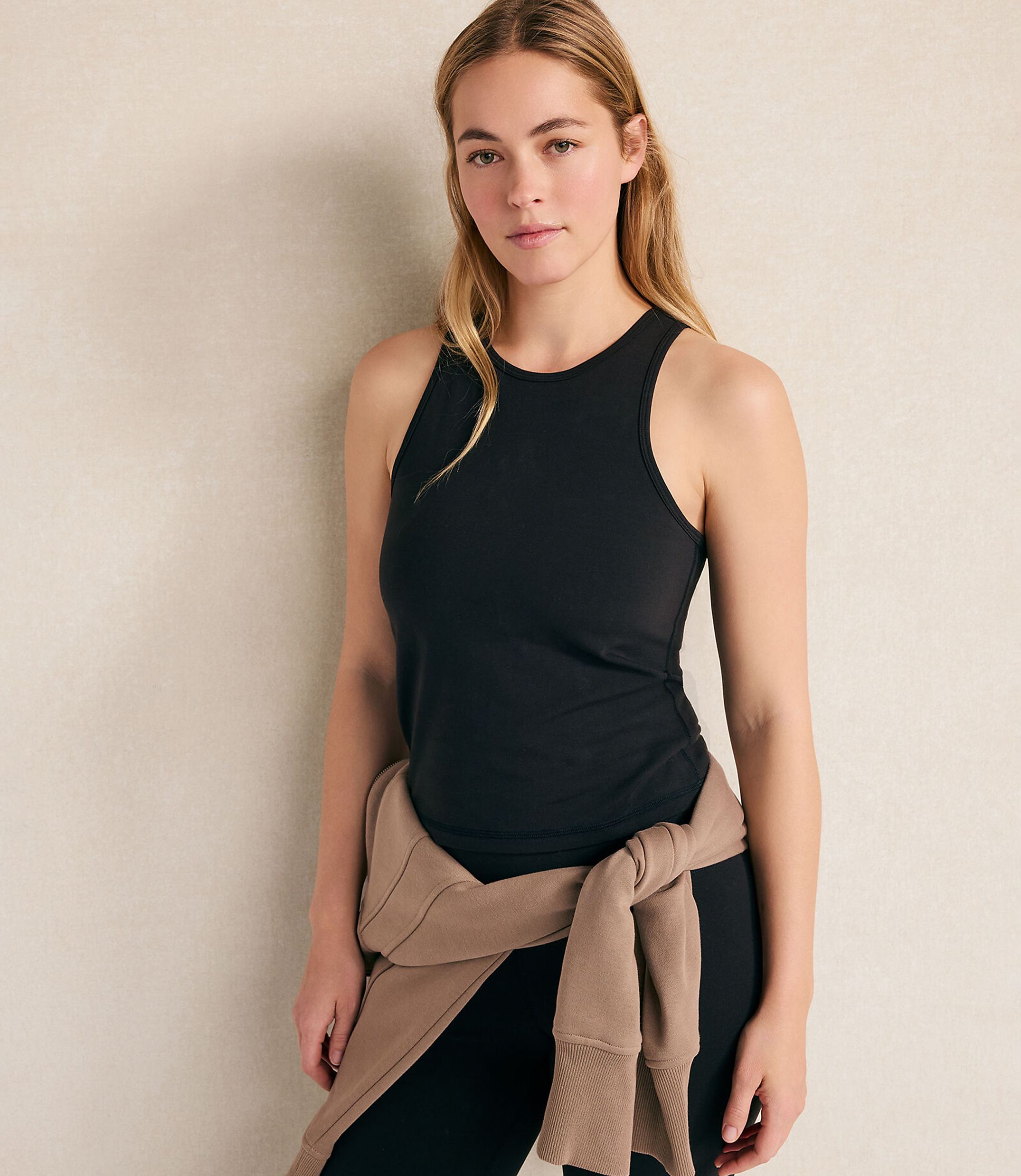 A woman wearing a black tank top and leggings with a tan sweater tied around her waist stands against a plain, light-colored wall.