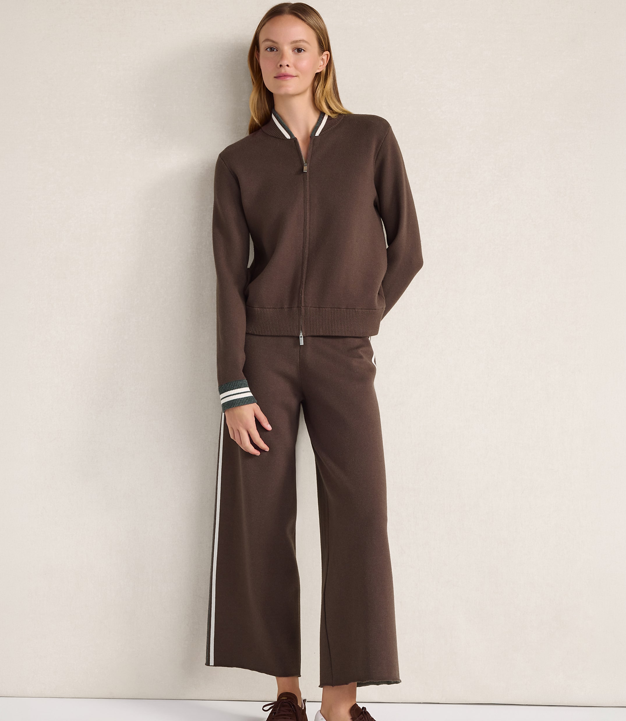 A woman wearing a brown zip-up jacket and matching wide-leg trousers stands against a plain, light-colored wall.
