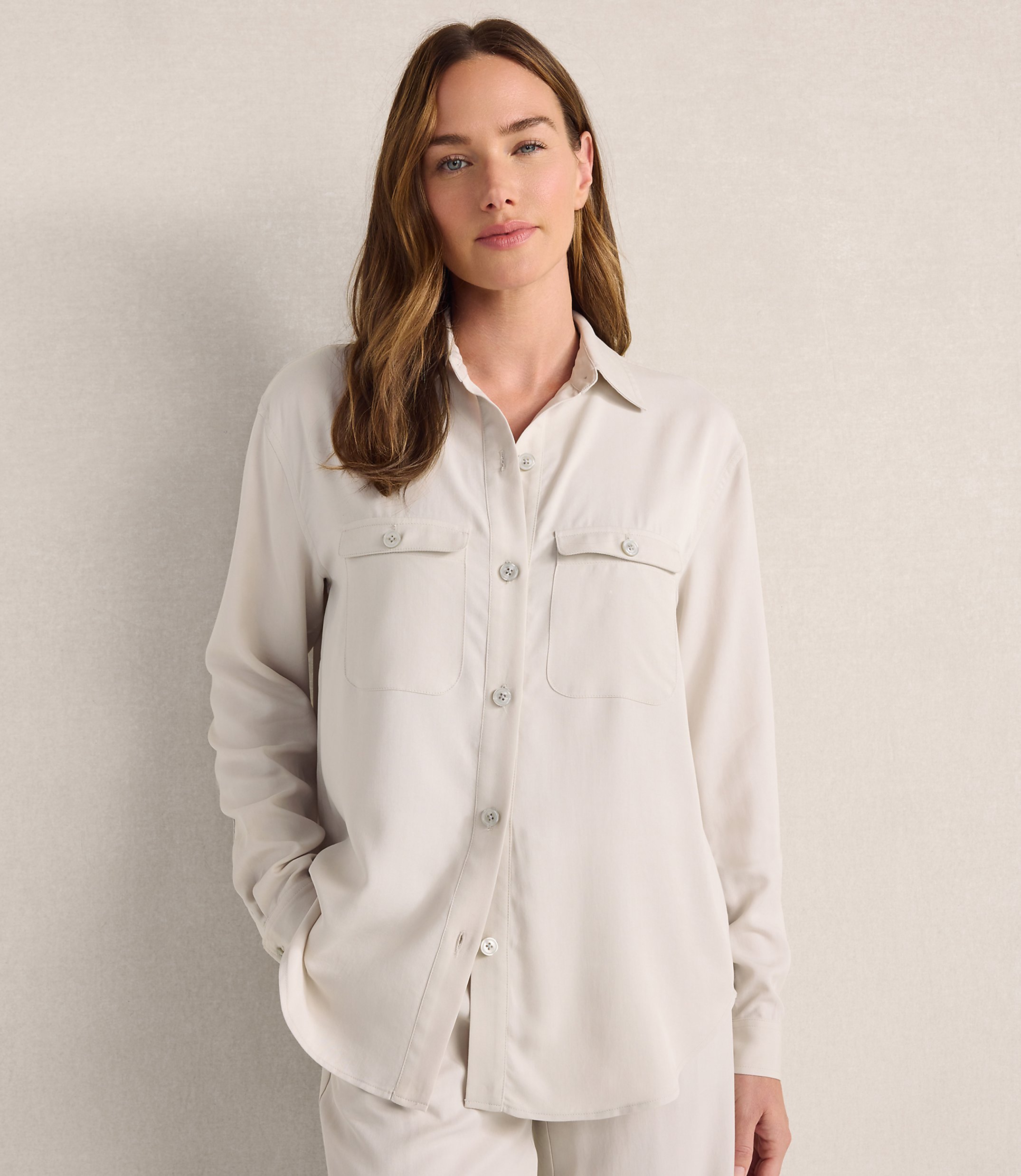 A woman wearing a light beige button-down shirt and matching pants stands against a plain, neutral-toned wall.